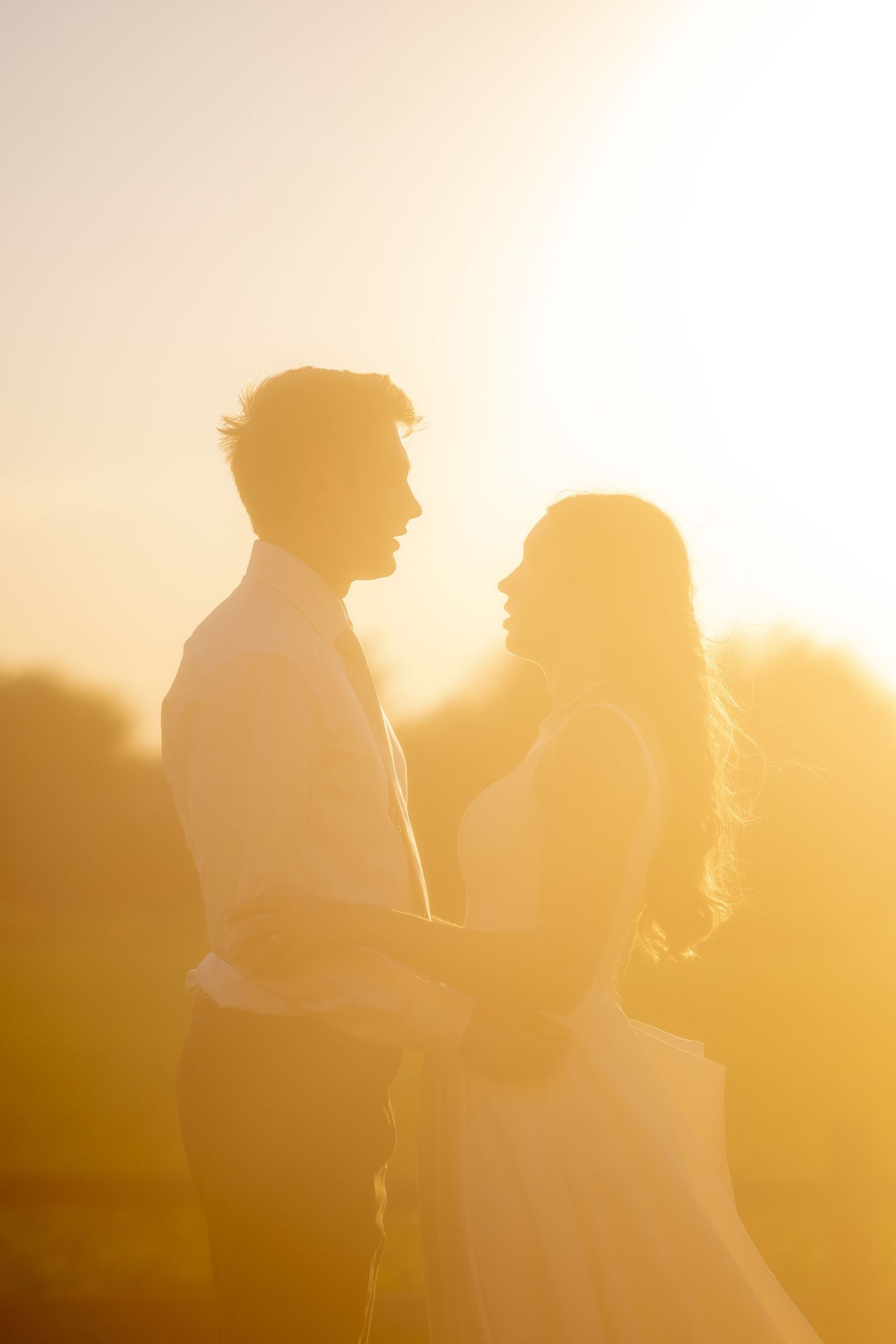 Romantic couple silhouetted against golden sunset light, embracing and gazing at each other outdoors.