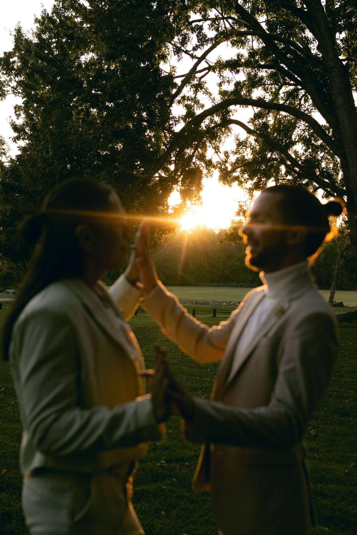 Two figures silhouetted against a golden sunset through trees share a tender moment in formal attire.