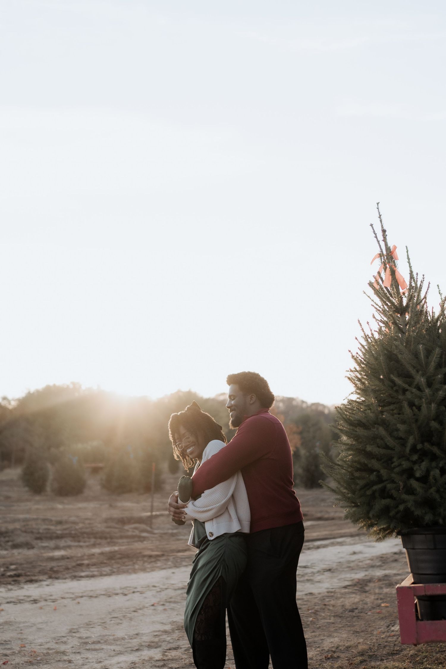Couple embracing and laughing at a Christmas tree farm during golden hour sunset.