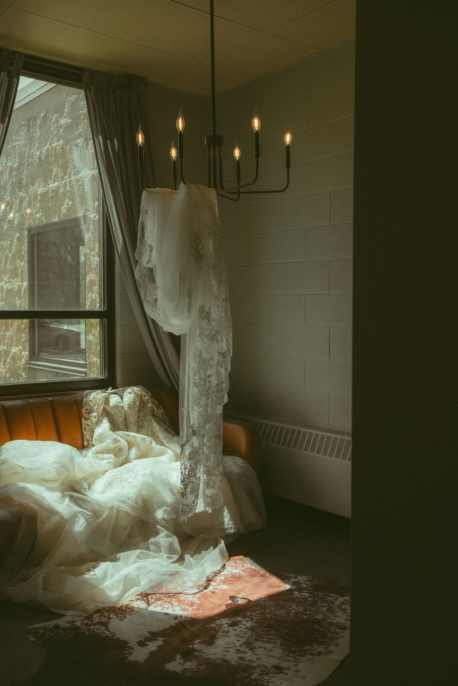 A white lace wedding dress hanging from a chandelier in a moody, dimly lit room with draped curtains and natural light.