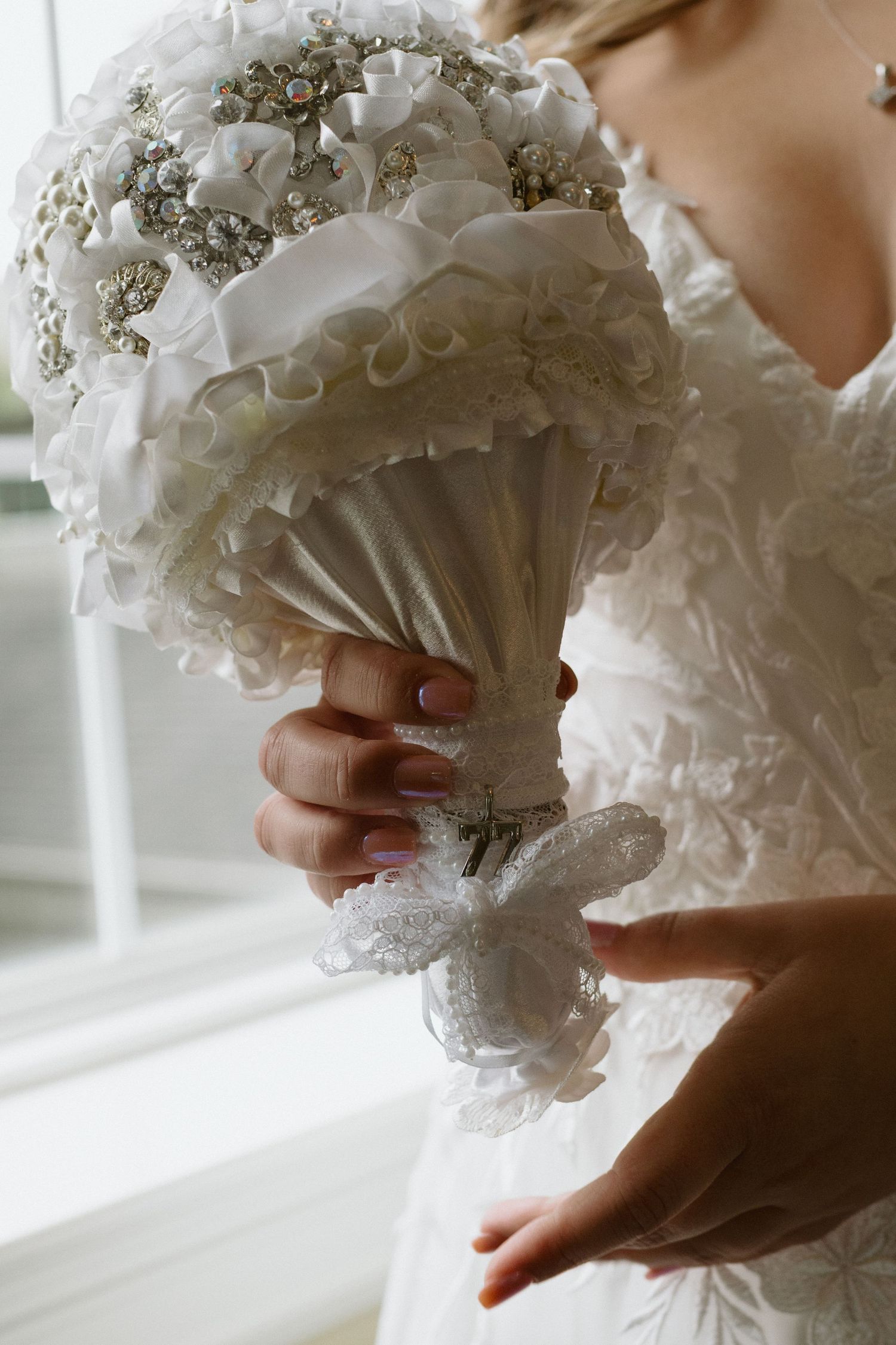 Elegant white bridal bouquet with crystal embellishments held against a lace wedding dress in soft natural light.