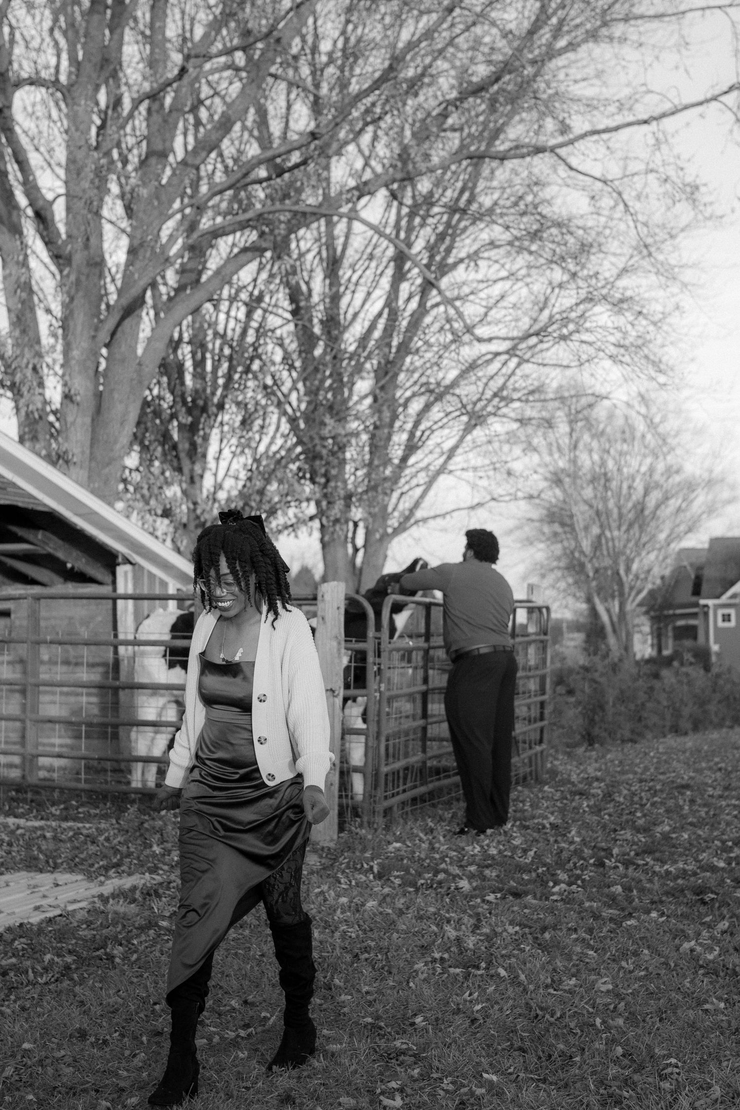 Black and white photo of a smiling woman in a white jacket and boots standing near a farm gate with a man in the background.