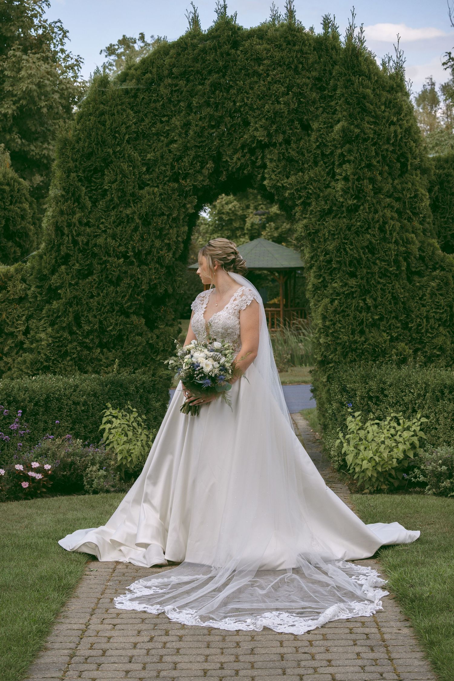 Bride in elegant white lace gown holding bouquet stands on garden path framed by sculpted evergreen hedges.