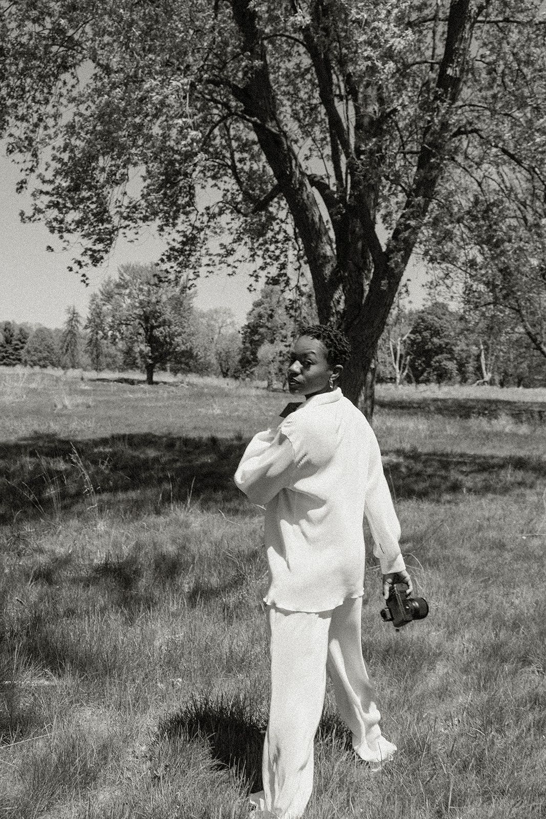 A person in all white clothing stands in a grassy field under trees, taking a photograph with a camera.