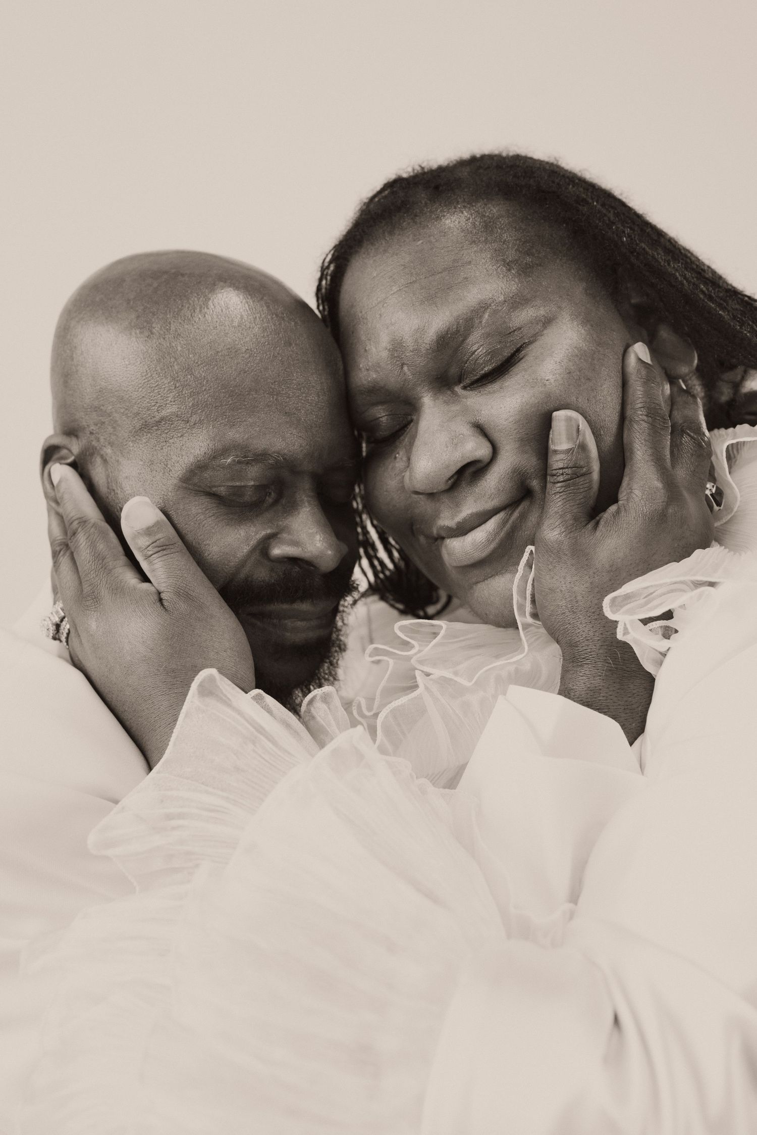 Black and white photo of a loving couple in white clothing, tenderly embracing with eyes closed.