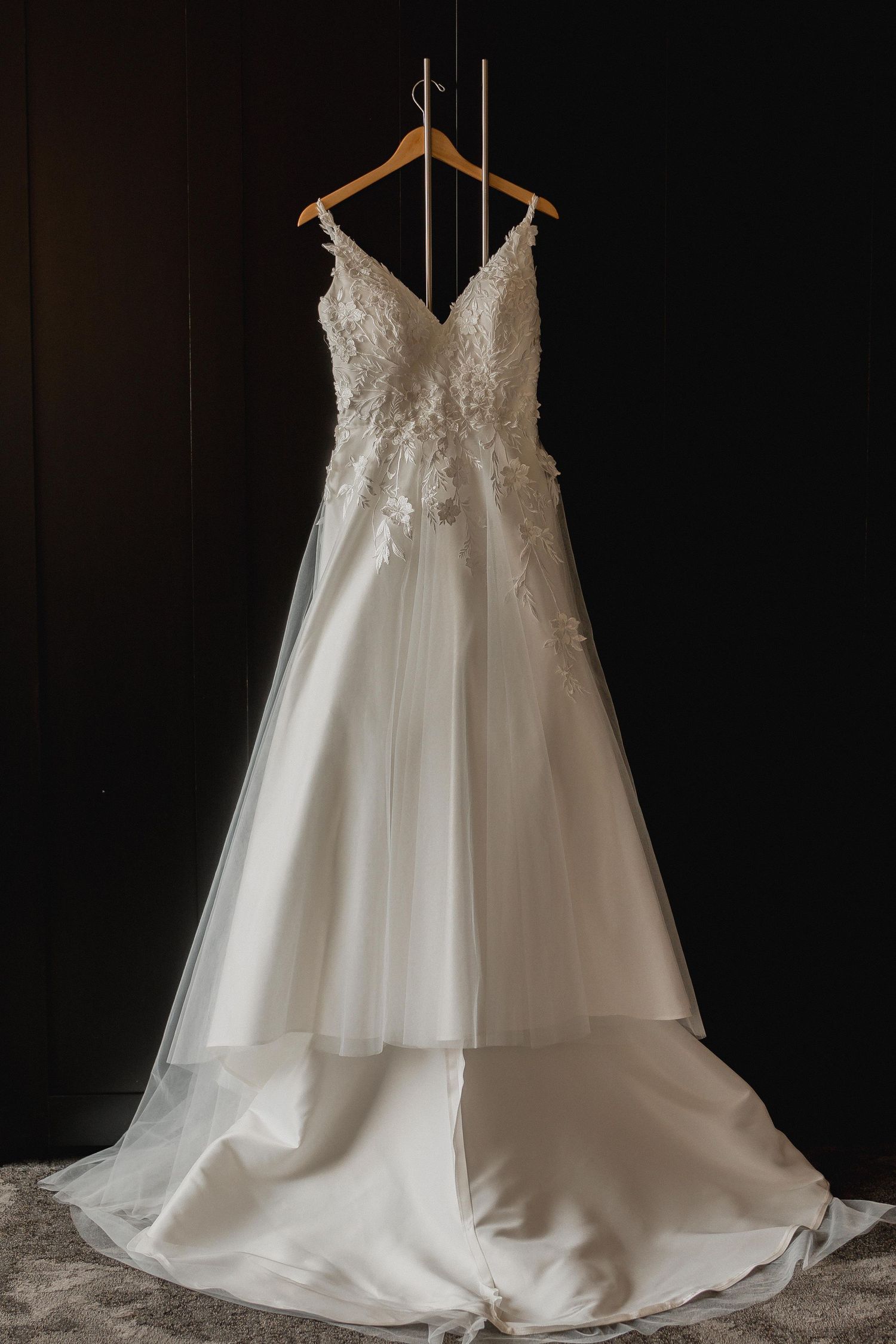 White wedding dress with lace bodice and flowing tulle skirt displayed on wooden hanger against dark background.