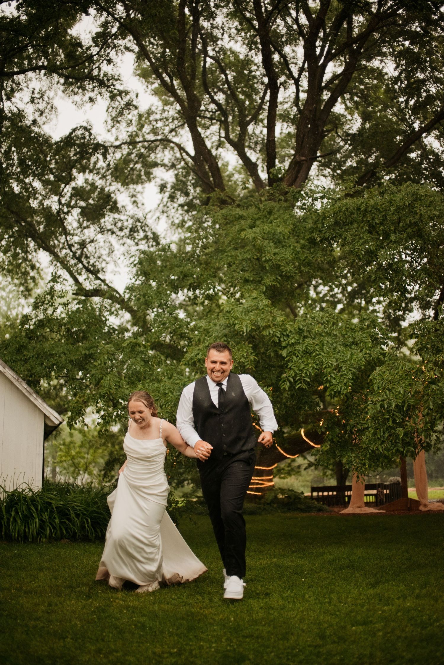 A joyful bride and groom run hand-in-hand across a lush green lawn at an outdoor wedding venue surrounded by trees.