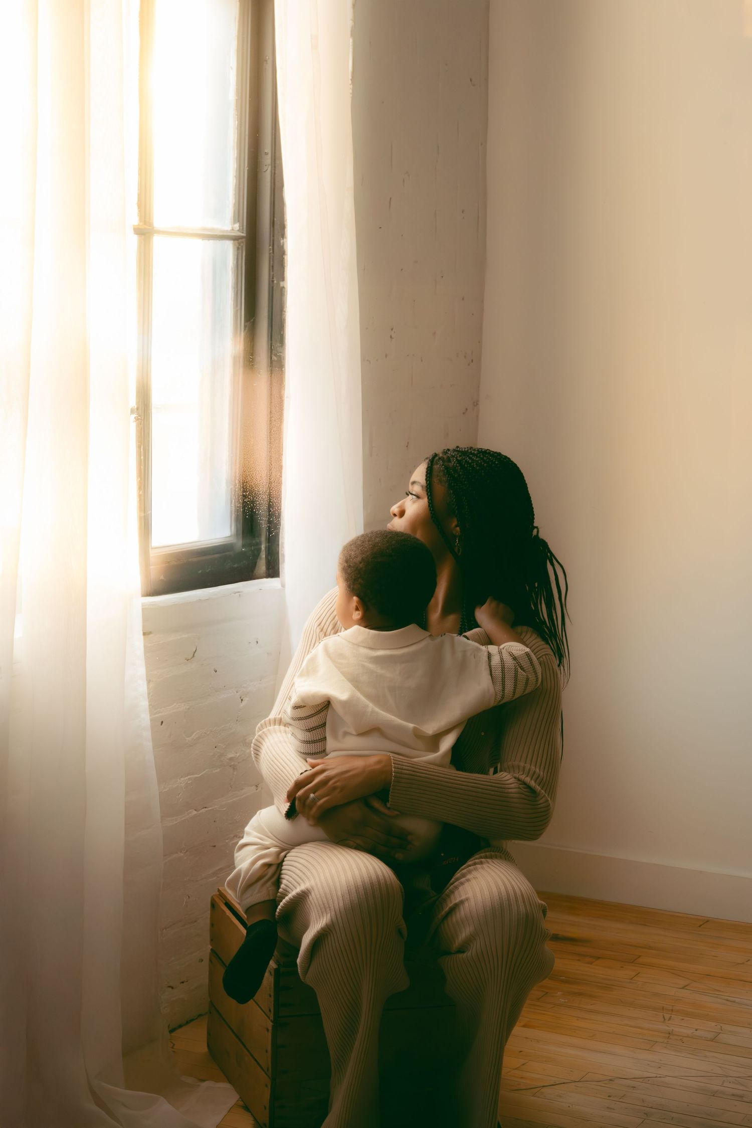 Two people share an intimate moment while sitting by a sunlit window in a minimalist room.