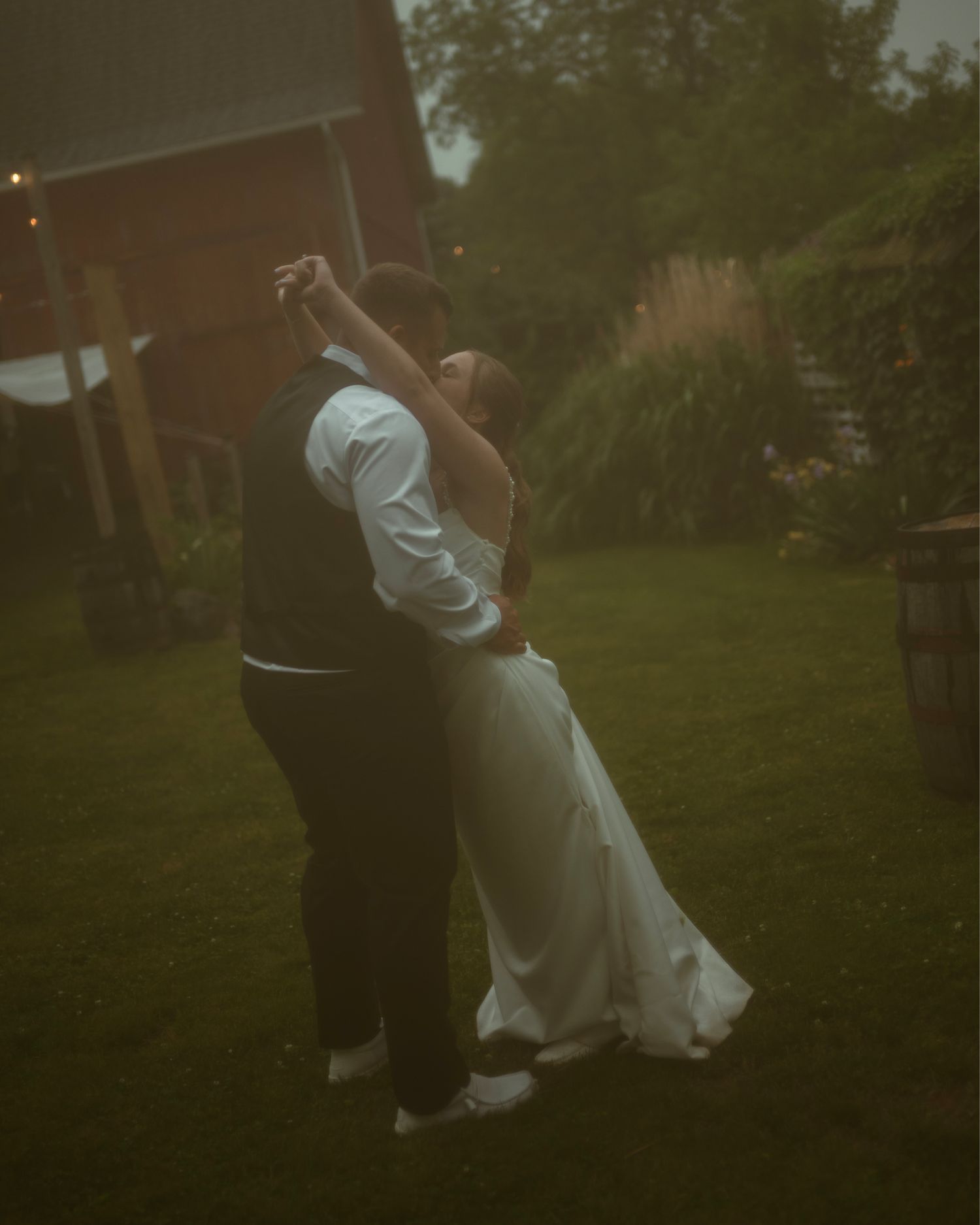 A bride and groom share a romantic first dance outdoors at dusk near a rustic red barn.