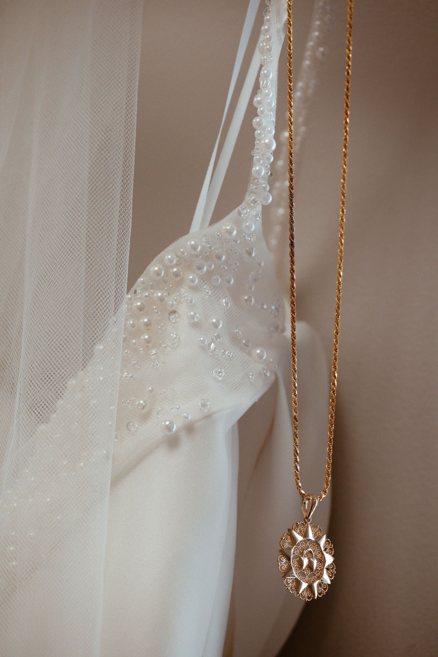 Delicate lace wedding veil hangs beside a dainty gold necklace with a floral pendant in soft natural light.
