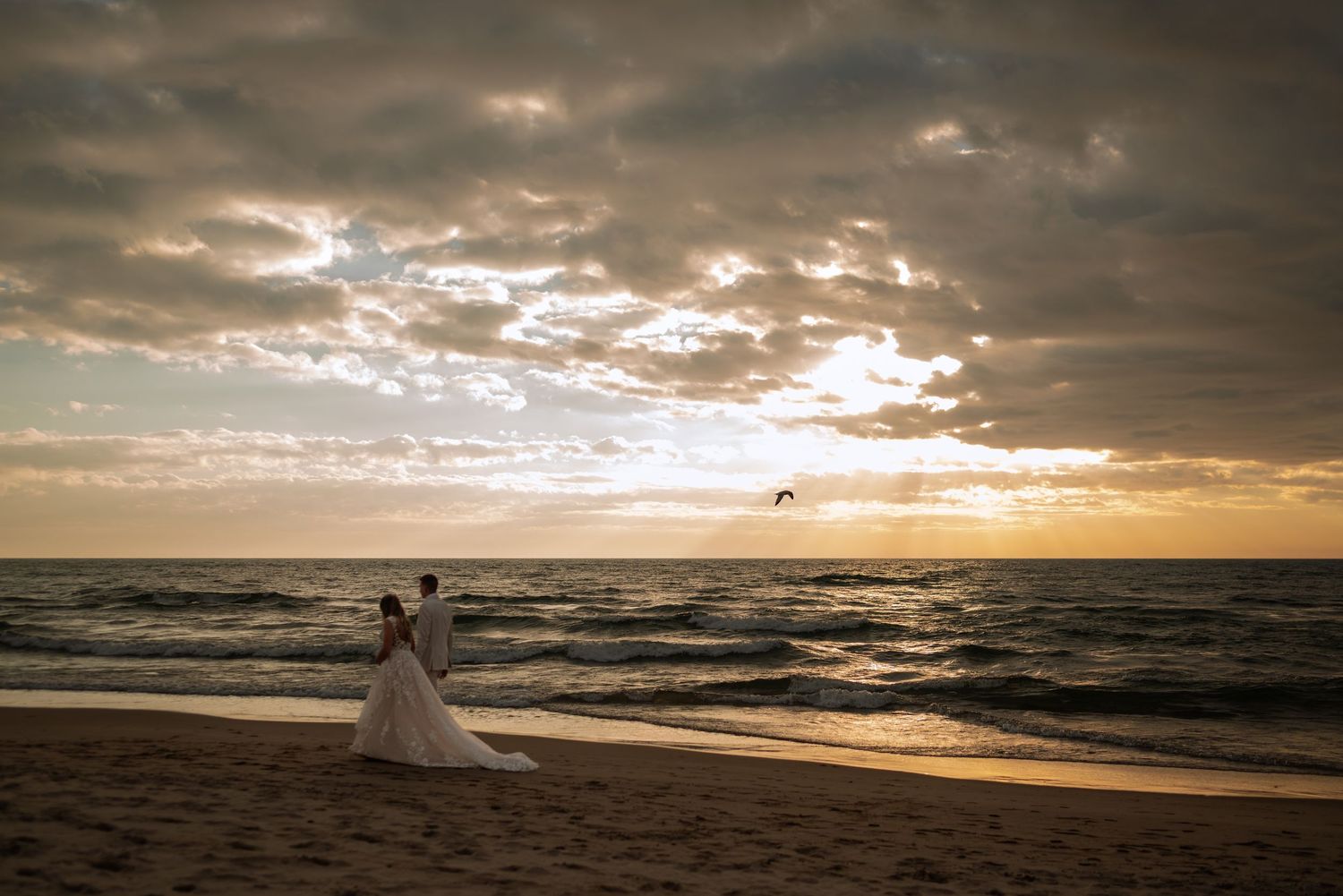 Bride and groom stand on a sandy beach at sunset, waves rolling in under a dramatic cloudy sky.