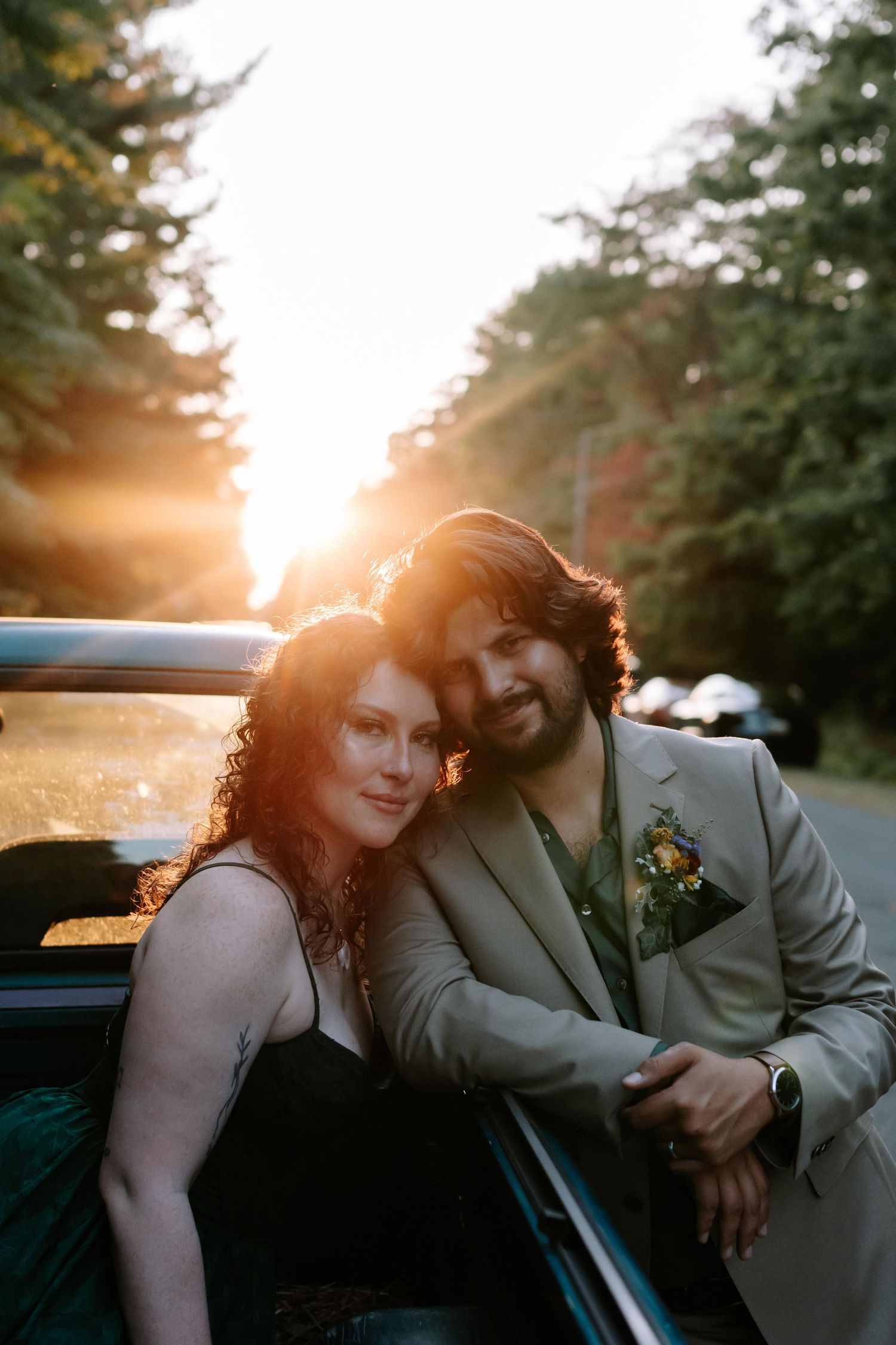 A couple leans together against a car during golden hour sunset with trees in the background.