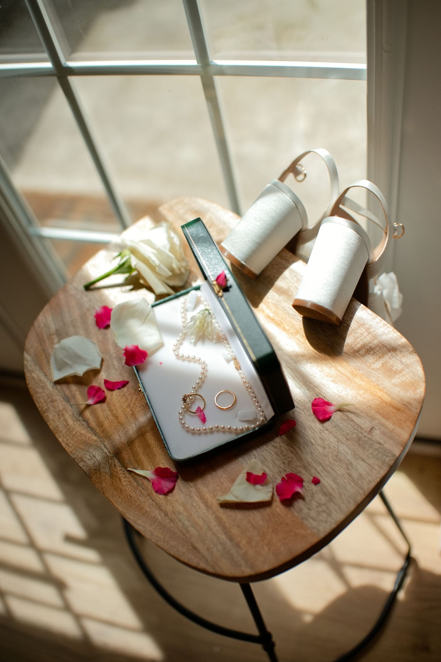 Bridal accessories on wooden table with pearl necklace, gold rings, white heels, and rose petals by a sunlit window.