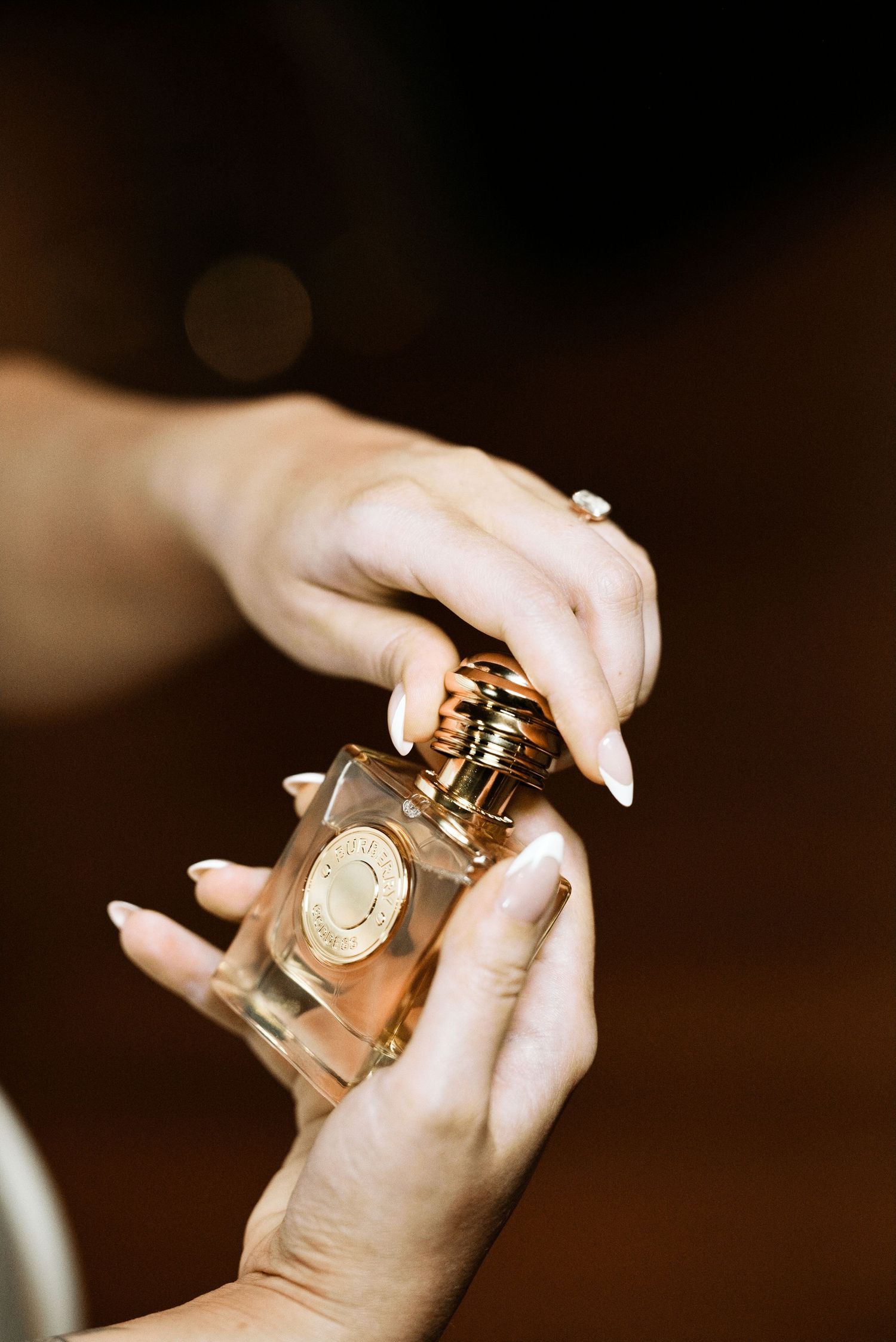 Close-up of elegant hands with manicured nails holding a luxury gold perfume bottle against dark background.