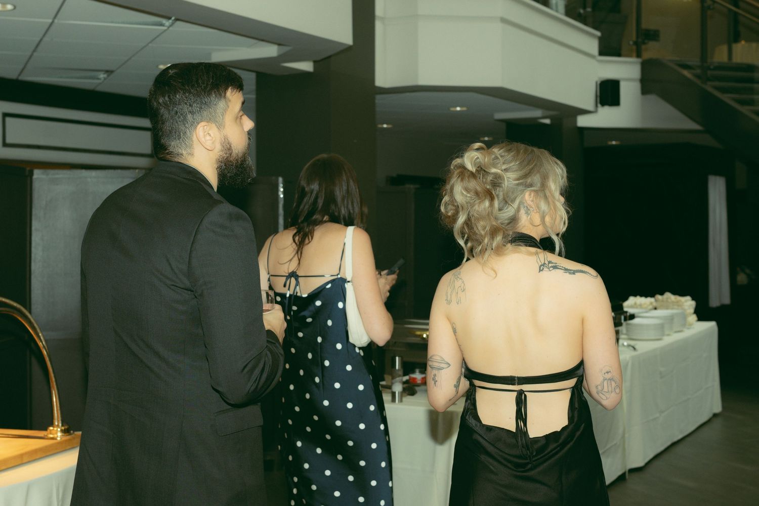 People mingling at a formal event wearing black formal attire and cocktail dresses in a dimly lit venue.