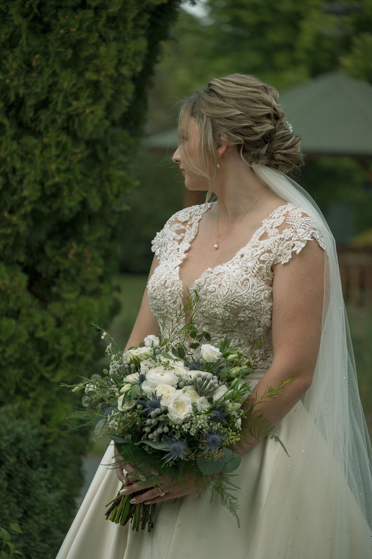 Side profile of bride in lace gown holding white and blue floral bouquet against garden backdrop.