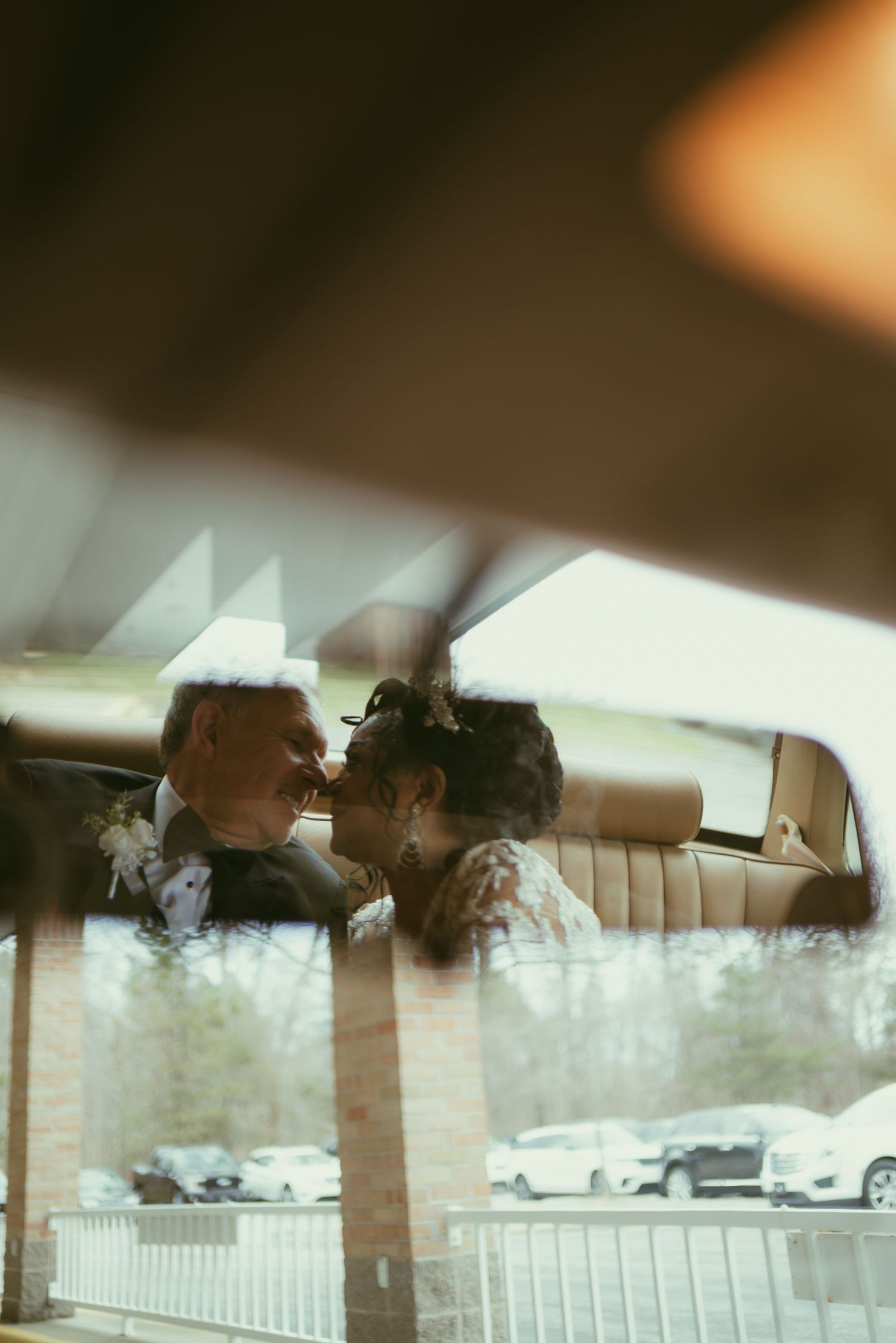 Couple sharing a romantic kiss in the backseat of a vintage car, captured through the window on their wedding day.