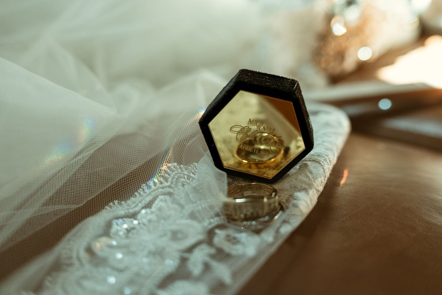 Gold wedding rings displayed in a hexagonal black velvet box with bridal veil and lace details in soft sunlight.