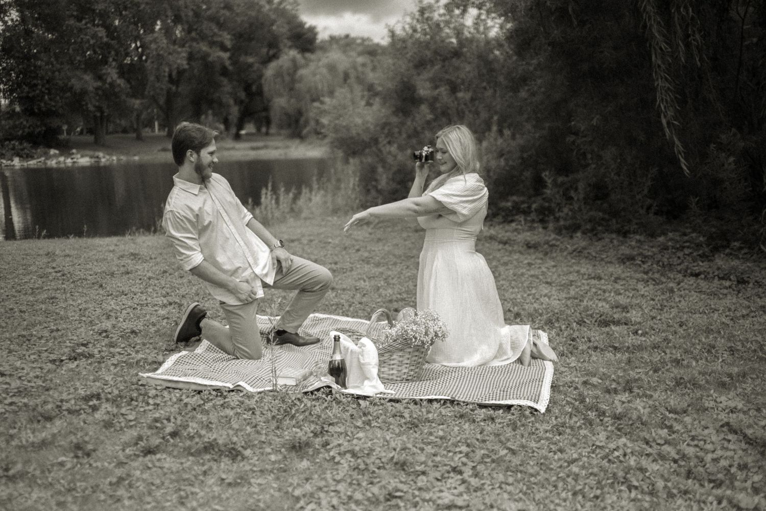 Black and white photo of a man on one knee proposing to a woman during a romantic outdoor picnic by a lake.
