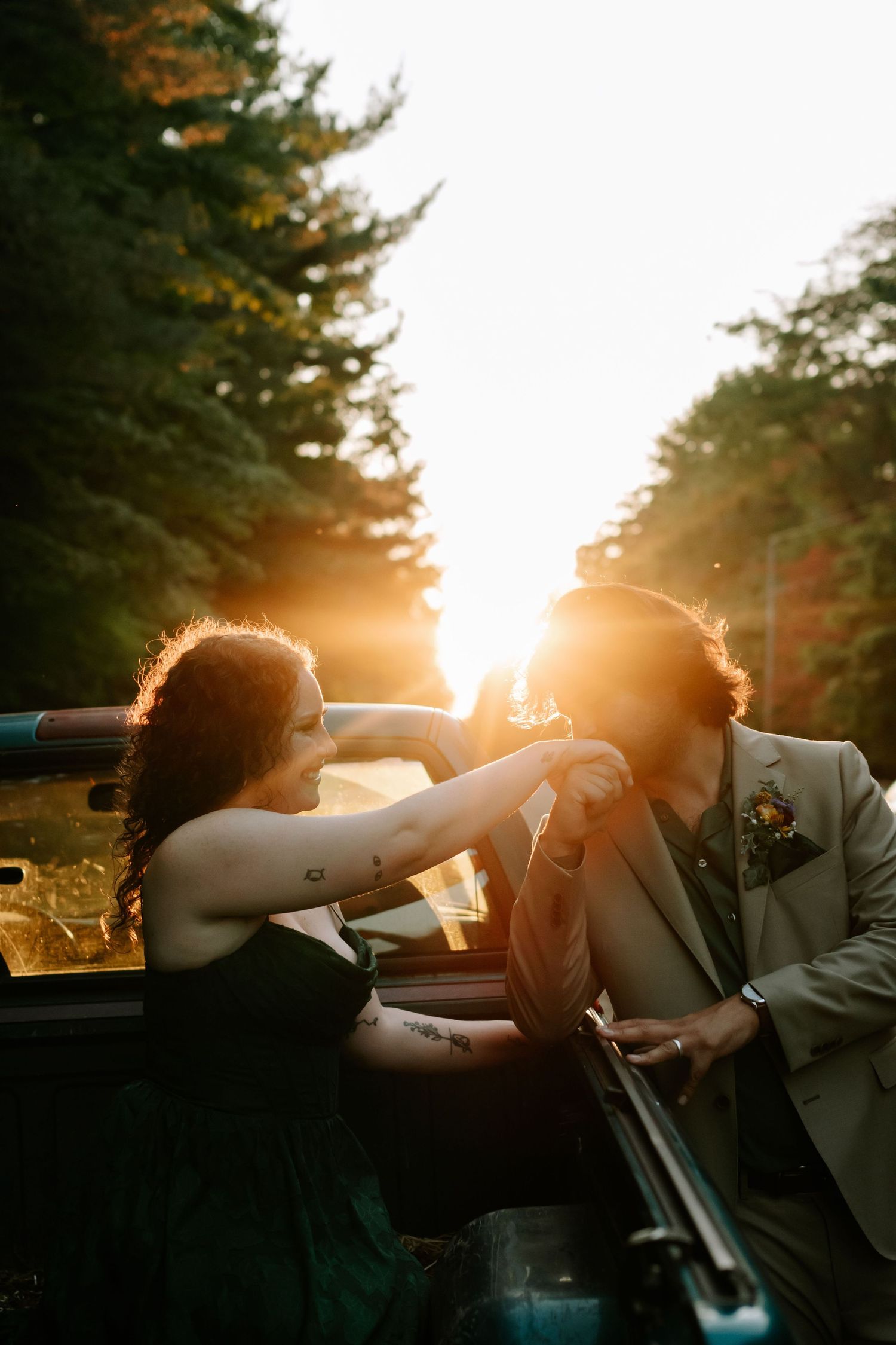 Silhouetted couple in a convertible car at sunset creating a romantic backlit scene through pine trees.