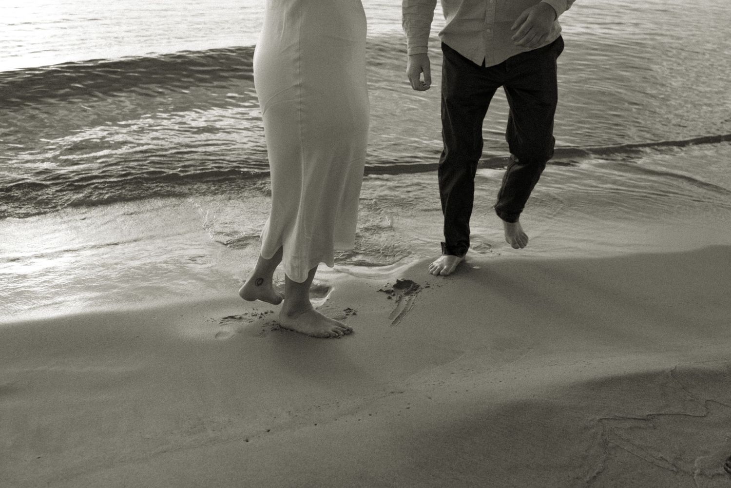 Black and white sequence showing couple's feet walking together on beach shoreline at sunset.
