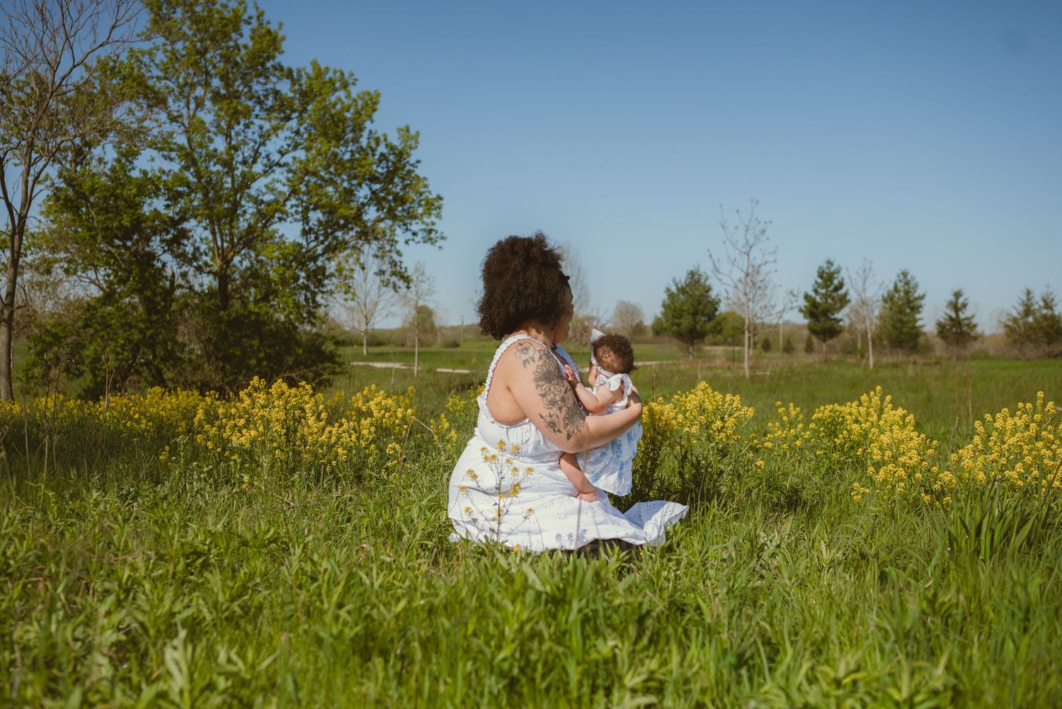 A mother sits in a lush green meadow with yellow wildflowers, tenderly holding her baby on a sunny day.