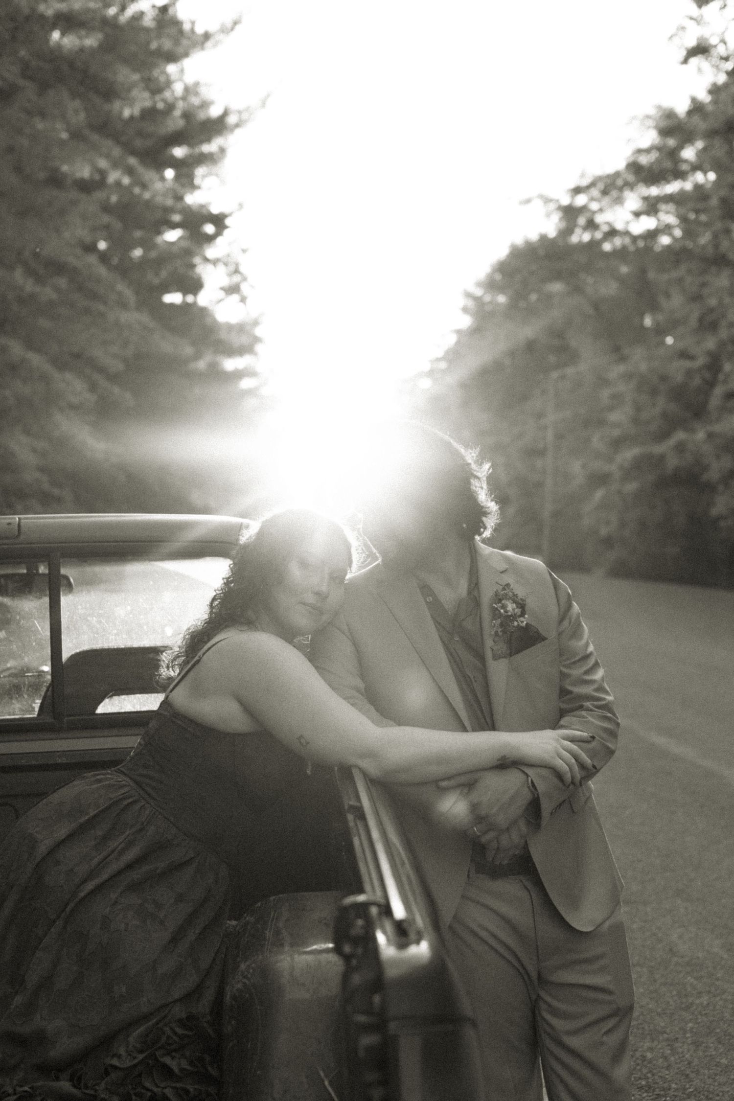 Four black and white photos showing a couple in romantic poses against bright sunlight near a vintage car on a rural road.