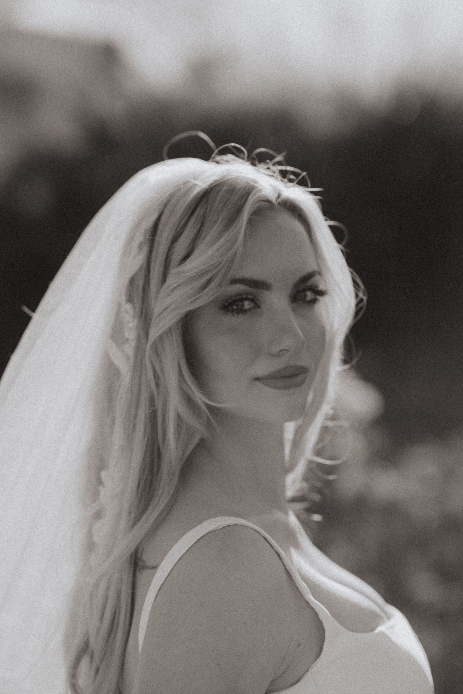 Black and white portrait of a bride with long wavy hair and a sheer veil, looking over her shoulder in soft natural light.