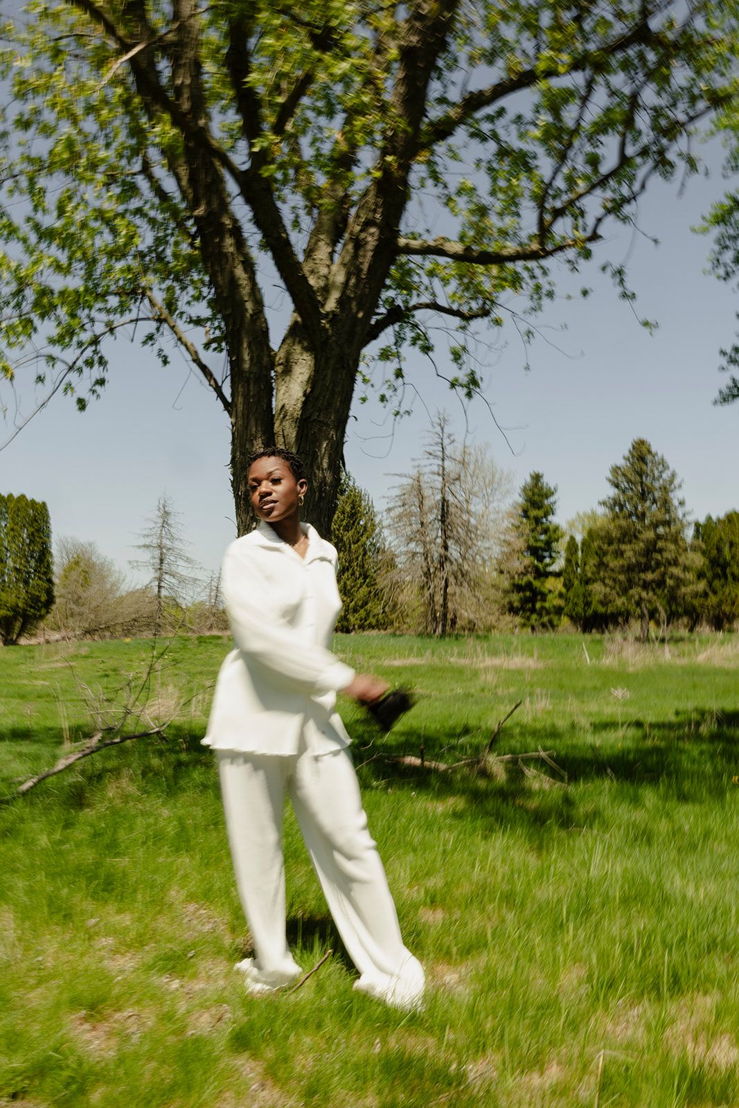 A person in a white outfit stands on green grass near a large tree on a sunny day.