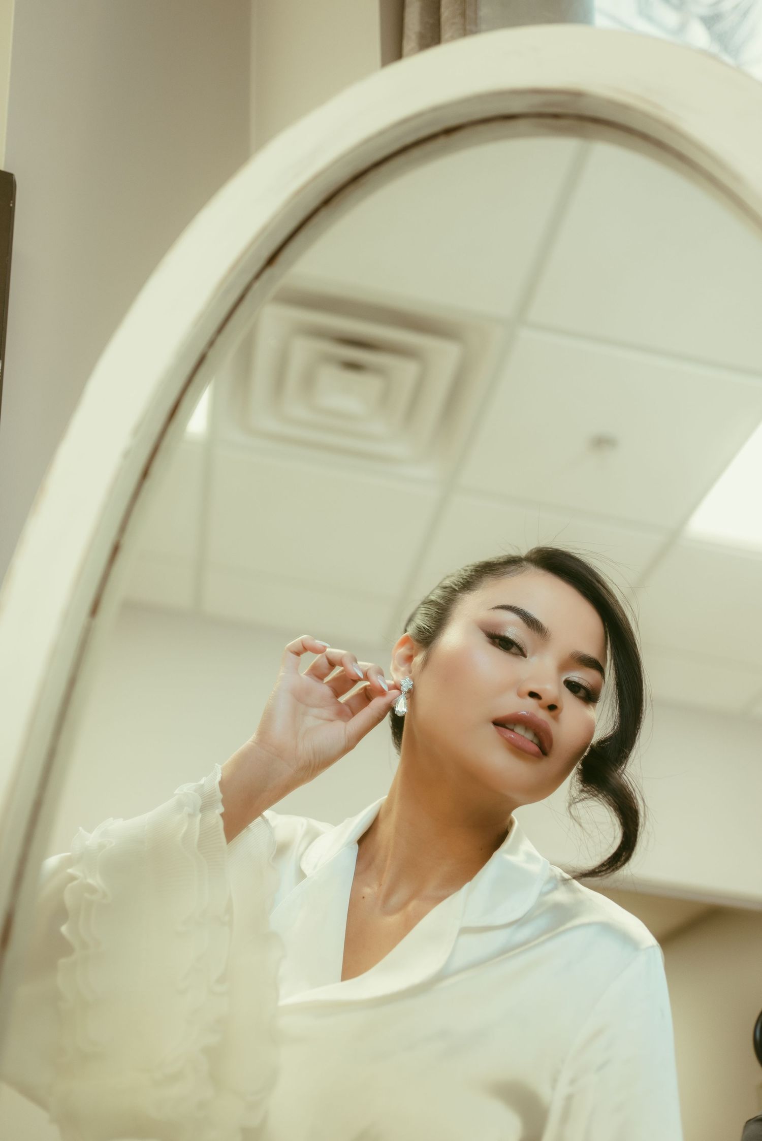 A bride in a white robe puts on earrings while looking in an arched mirror, preparing for her wedding day.