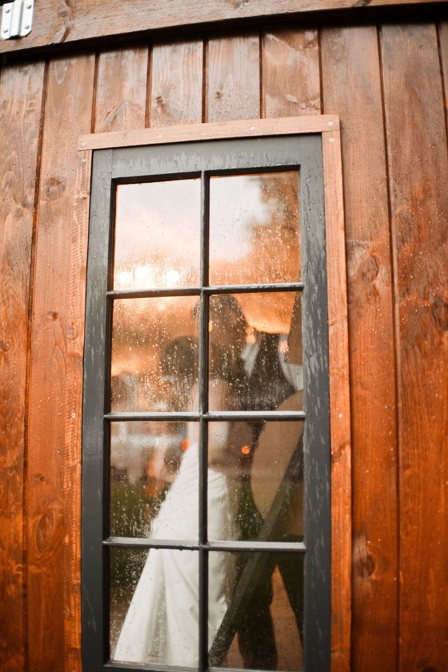 A couple shares a kiss behind a rain-speckled glass door of a rustic wooden barn at golden hour.