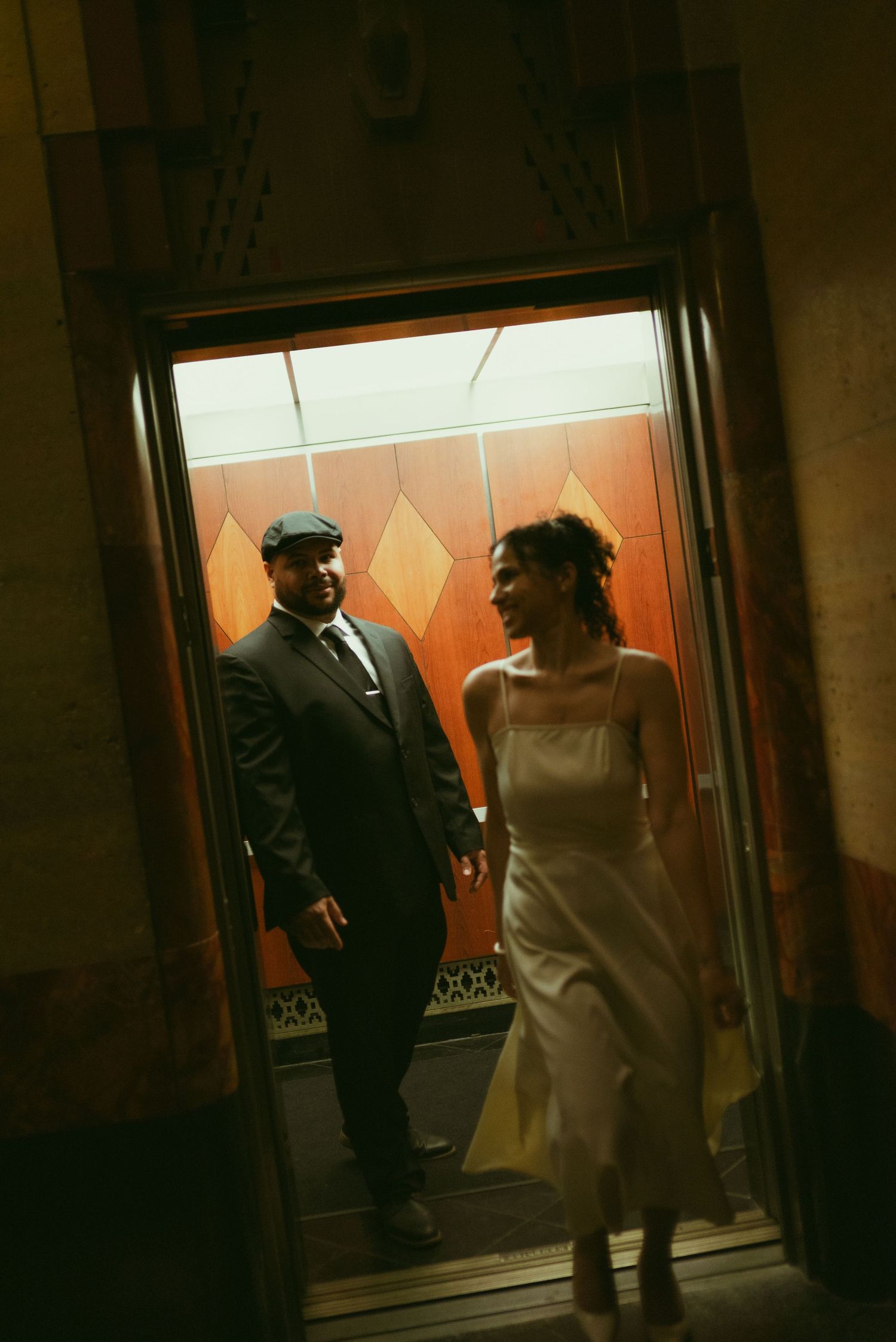 A couple in elegant attire shares a romantic moment stepping out of an ornate elevator with Art Deco decor.