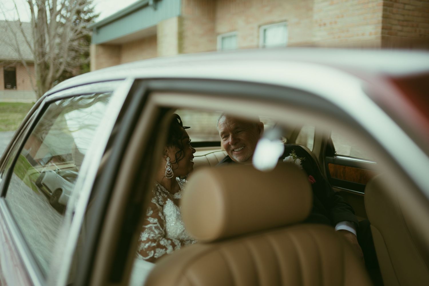 Bride and elderly man share a tender moment inside a vintage car with tan leather seats.