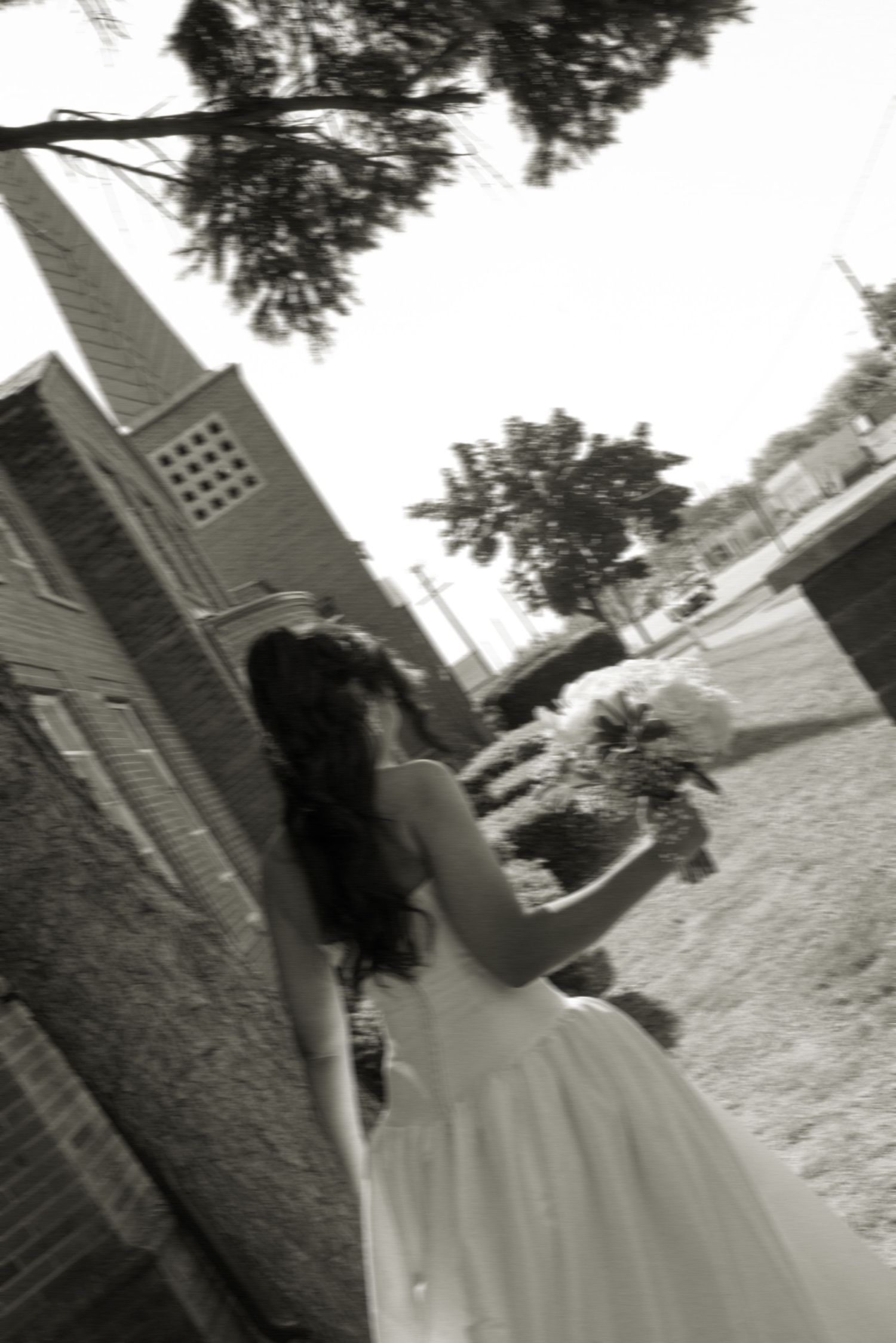 Black and white photo of a bride in a white gown holding a bouquet, walking toward a brick church.