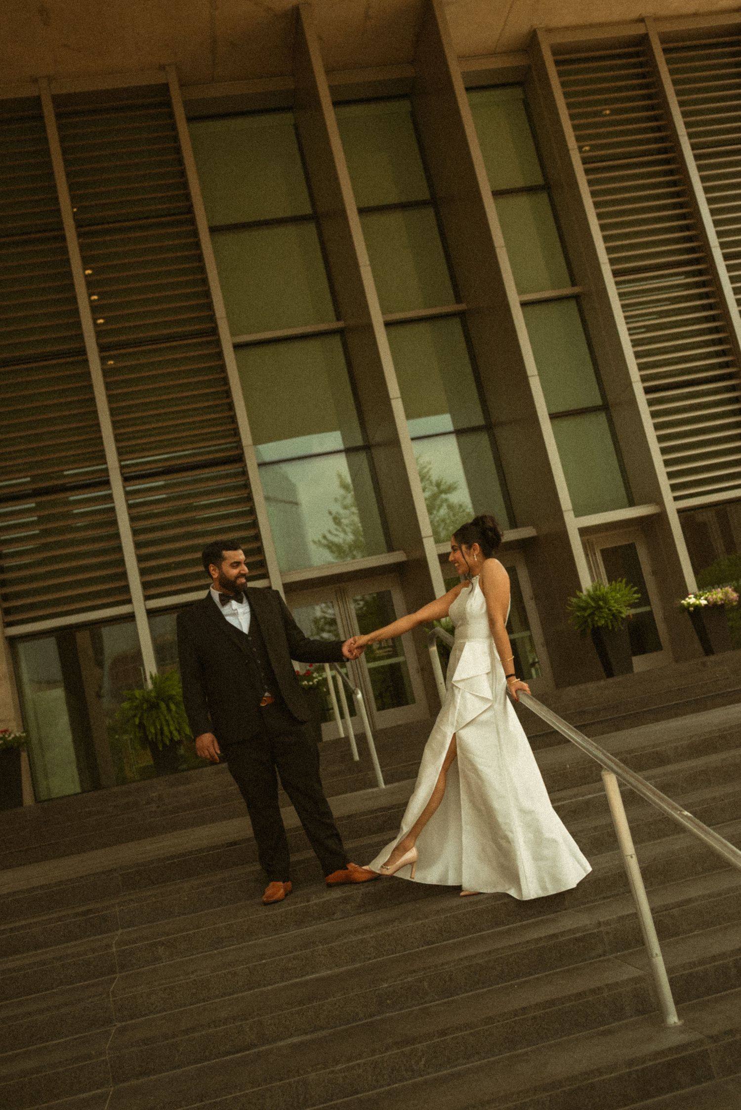 Bride and groom hold hands on steps outside modern building, bride in white gown, groom in black suit.