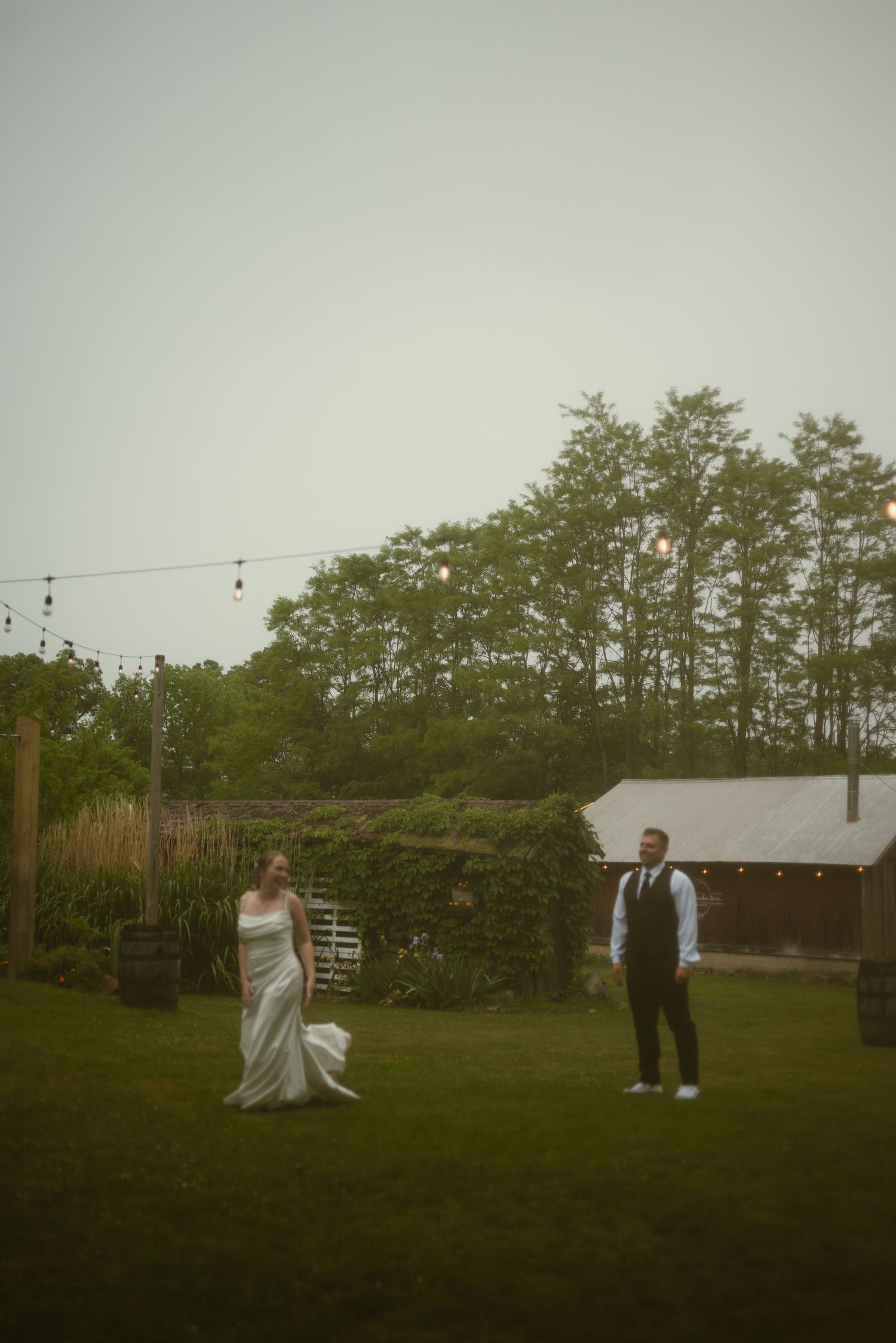 Bride in white gown and groom in vest share an outdoor moment on green lawn with string lights and barn at dusk.