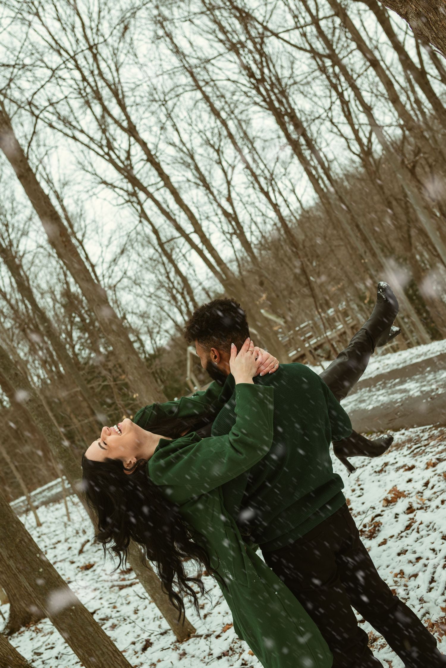 Couple dancing romantically in snowy winter forest, wearing matching green outfits, with snow falling around them.