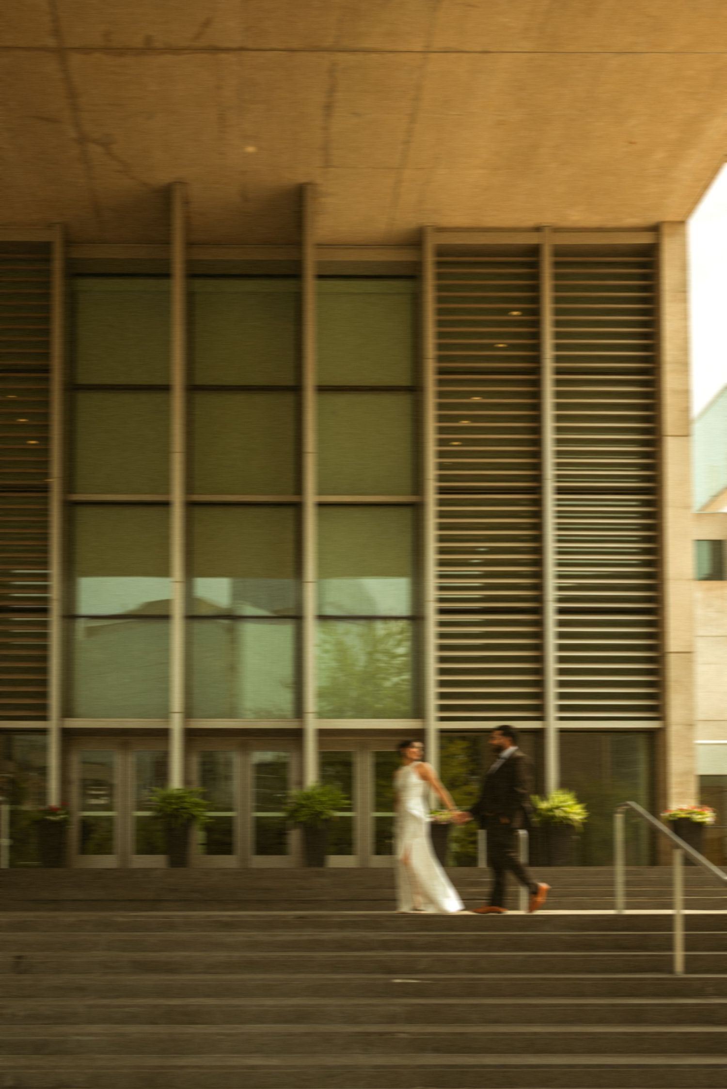 A bride and groom walk hand in hand on steps outside a modern concrete building with large glass windows.
