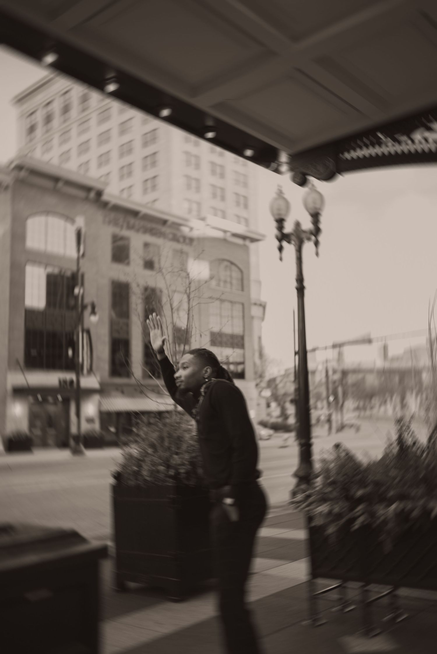 Black and white photo of a person raising their hand on a city street with a lamp post and buildings in the background.