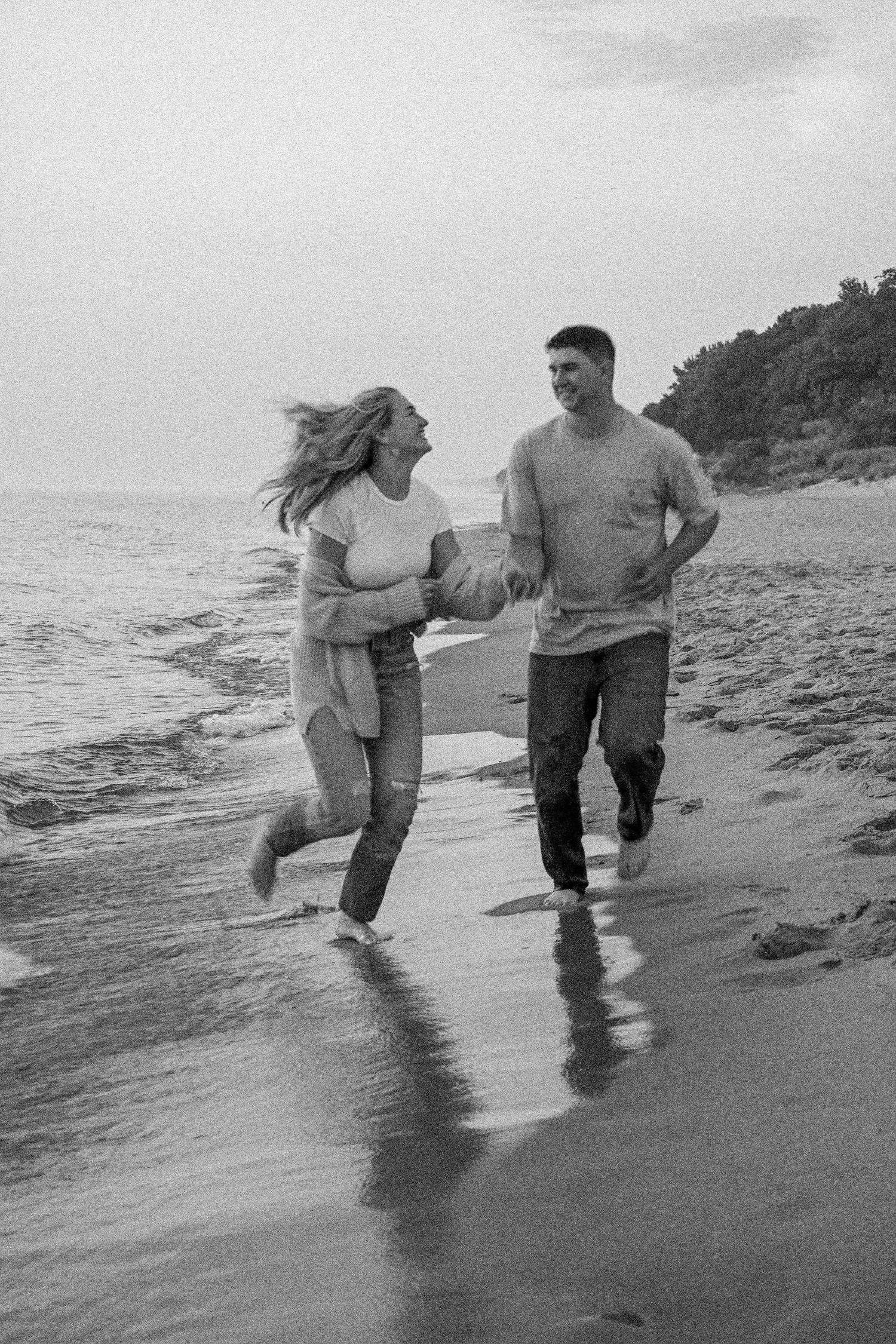 Black and white photo of a couple joyfully running together on a beach at sunset.