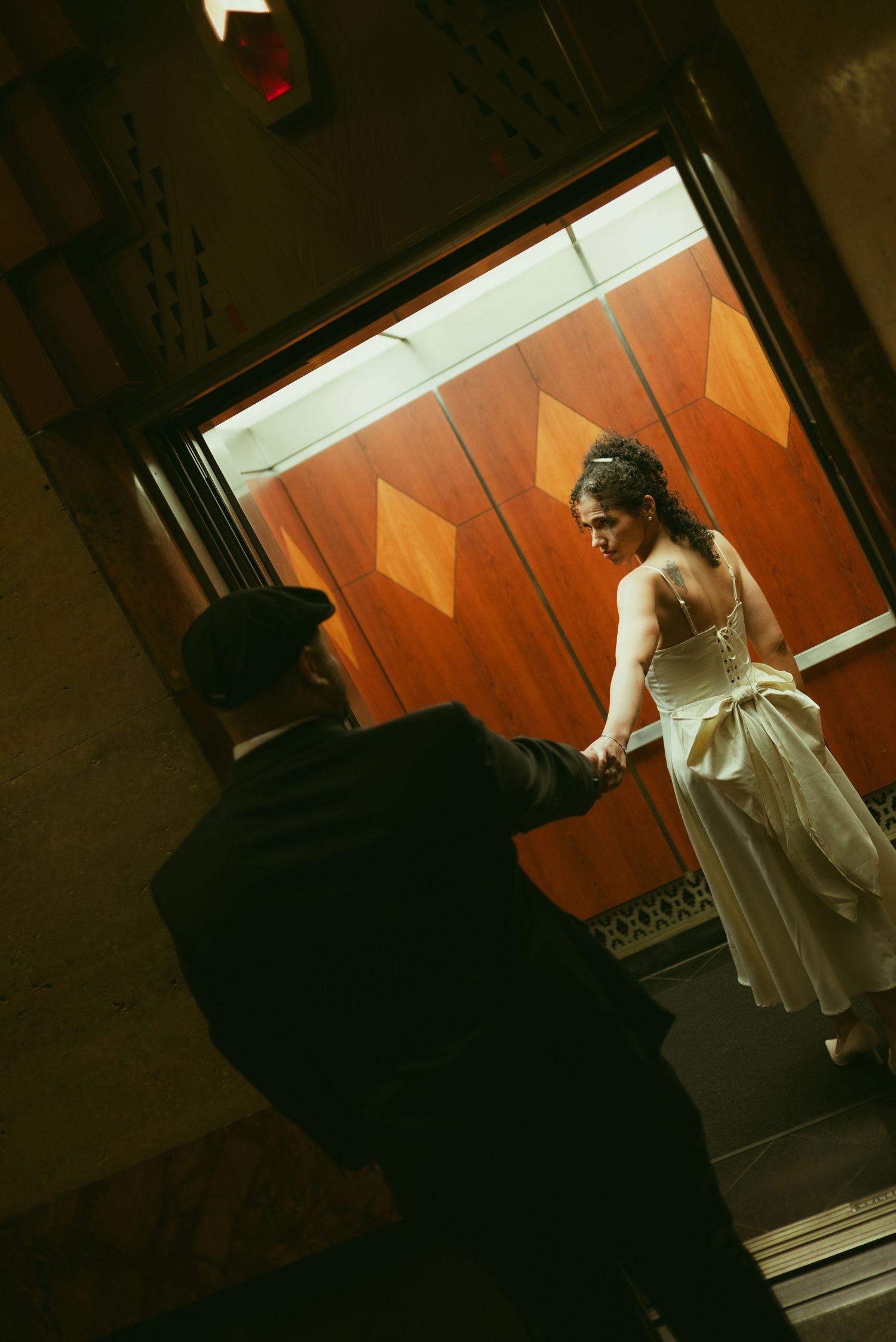 Bride in white gown holds groom's hand stepping into ornate wood-paneled elevator during wedding celebration.