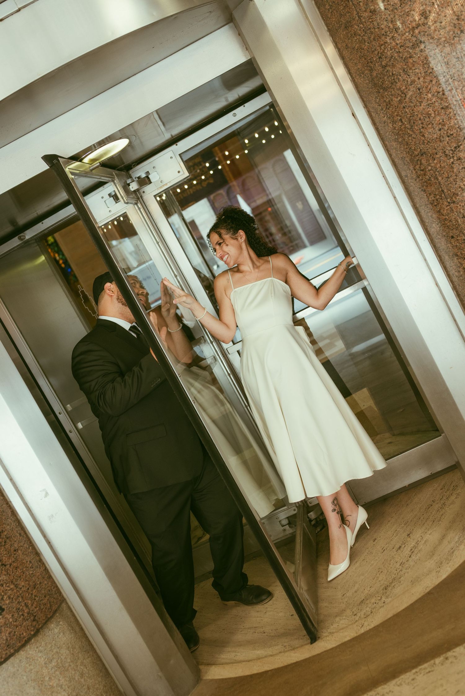 Bride in white dress and groom in black suit share a romantic moment inside a glass elevator.