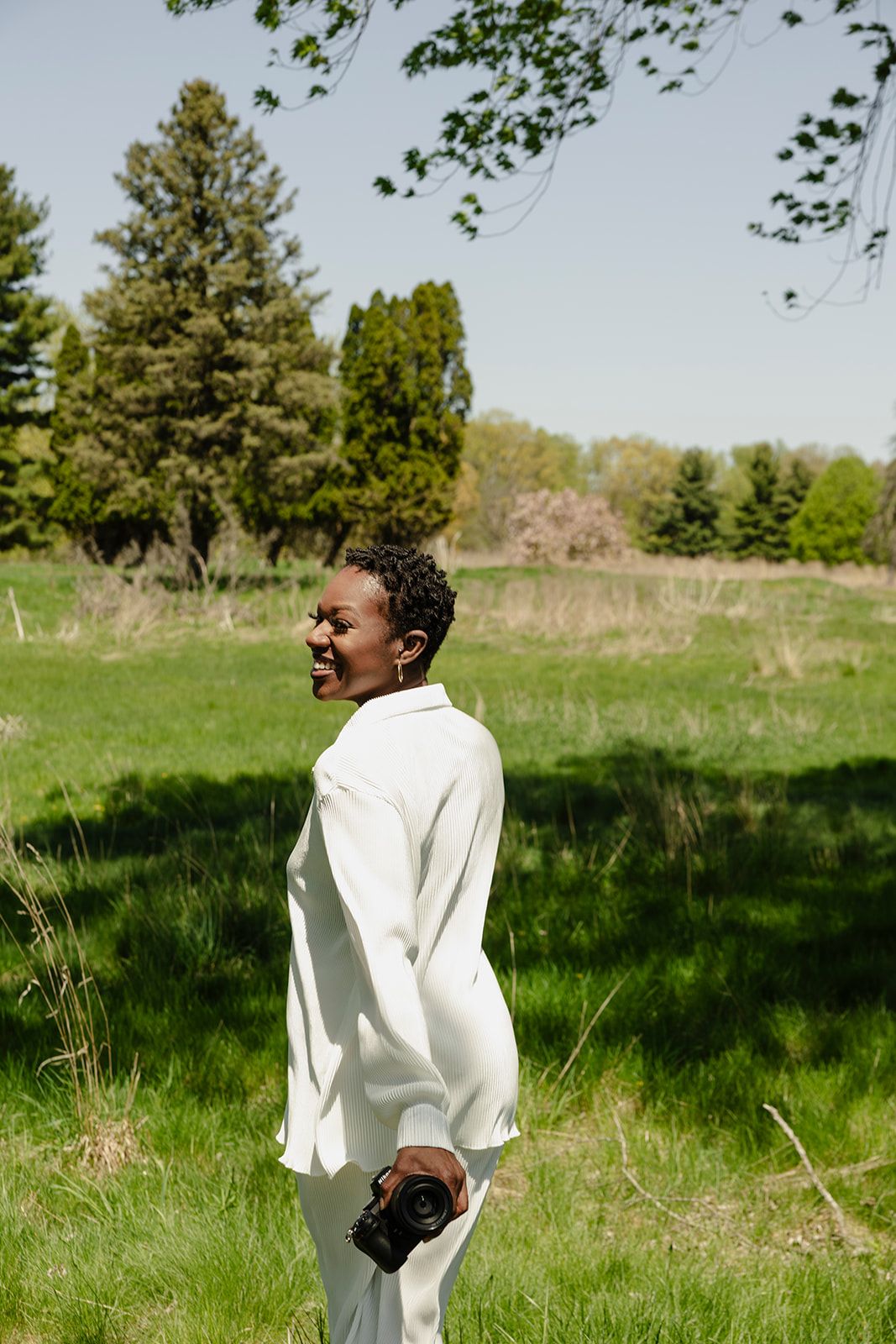 A smiling Black woman in a white outfit holds a camera while standing in a lush green field on a sunny day.