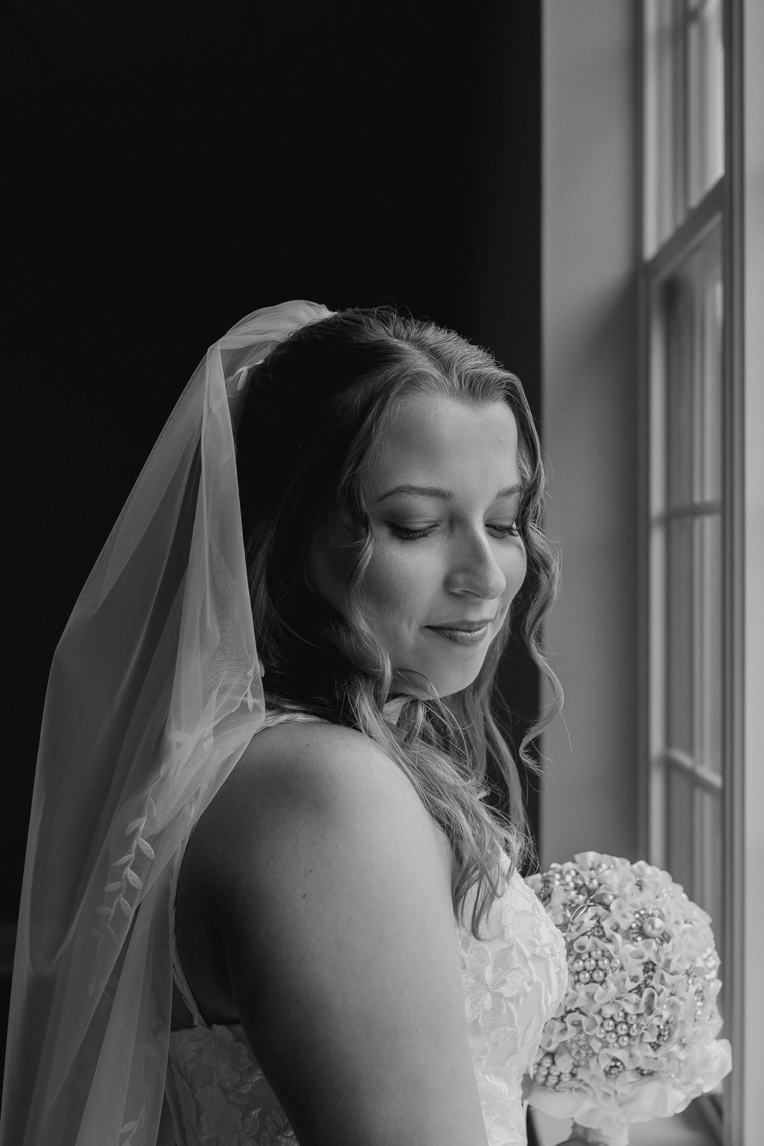 A bride in a white wedding dress and veil stands by a window holding a bouquet in black and white photography.