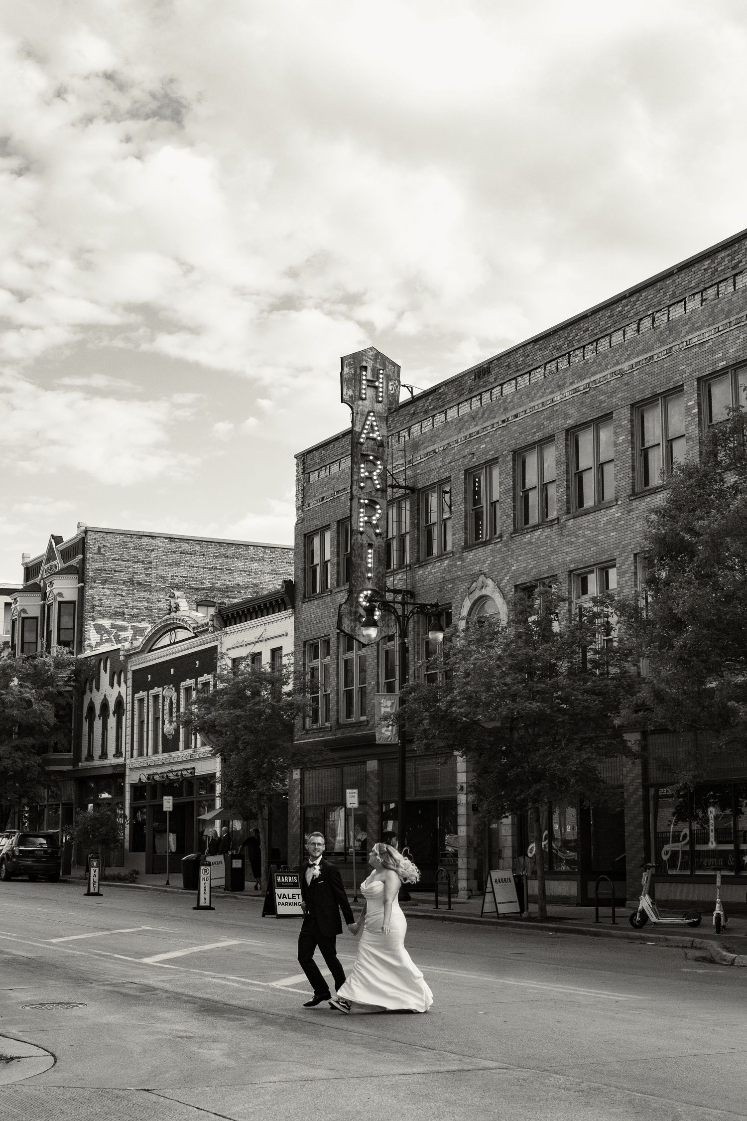 Black and white photo of a bride and groom crossing a street in front of a historic brick building with a Harris sign.