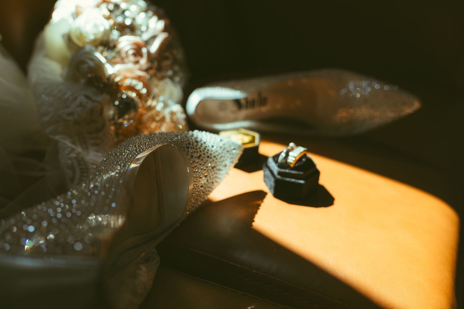 Close-up of wedding rings in a black box beside sparkly bridal heels and a bouquet in warm sunlight.