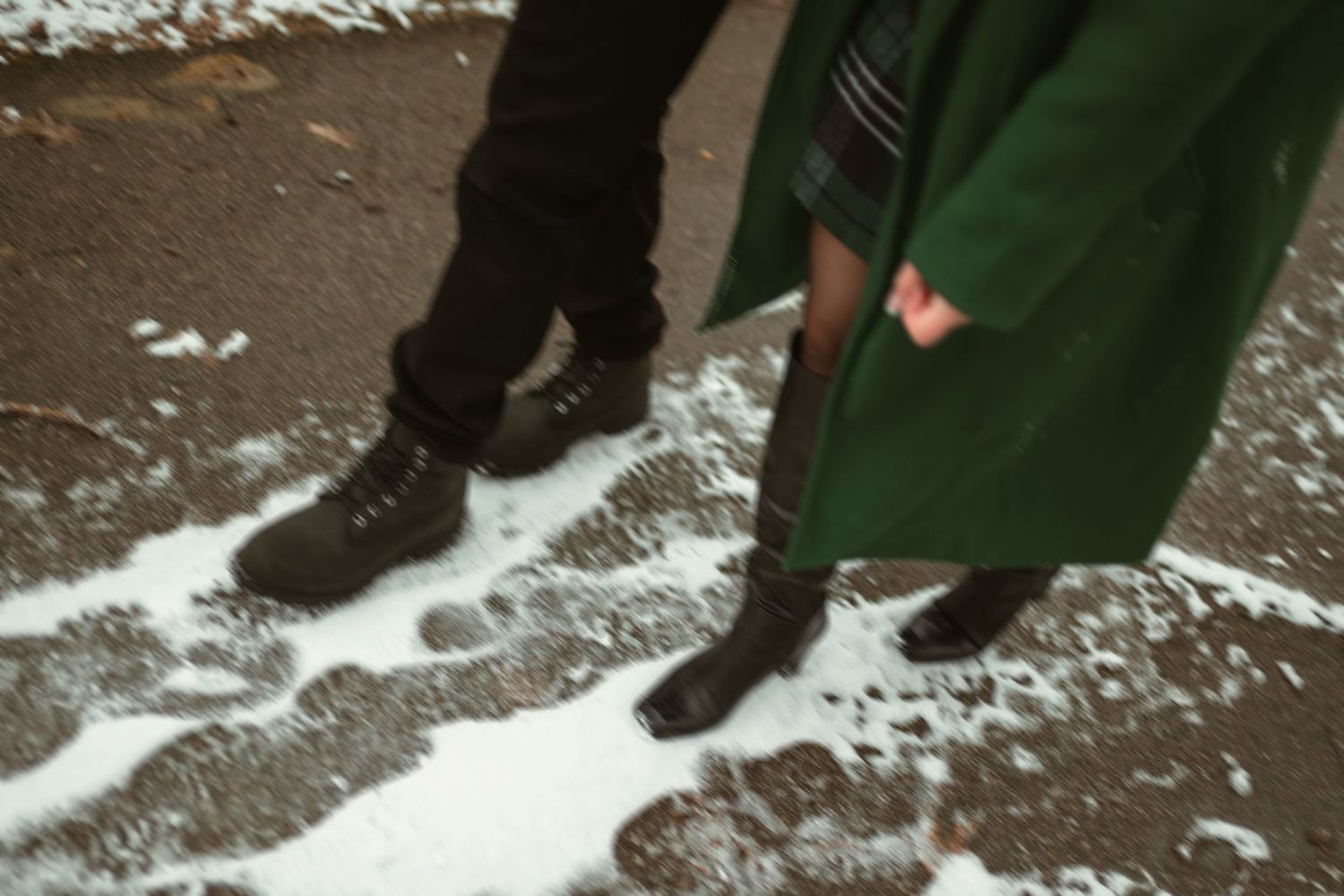 Two people wearing boots standing on a snowy wet pavement, one in a green coat and black boots.