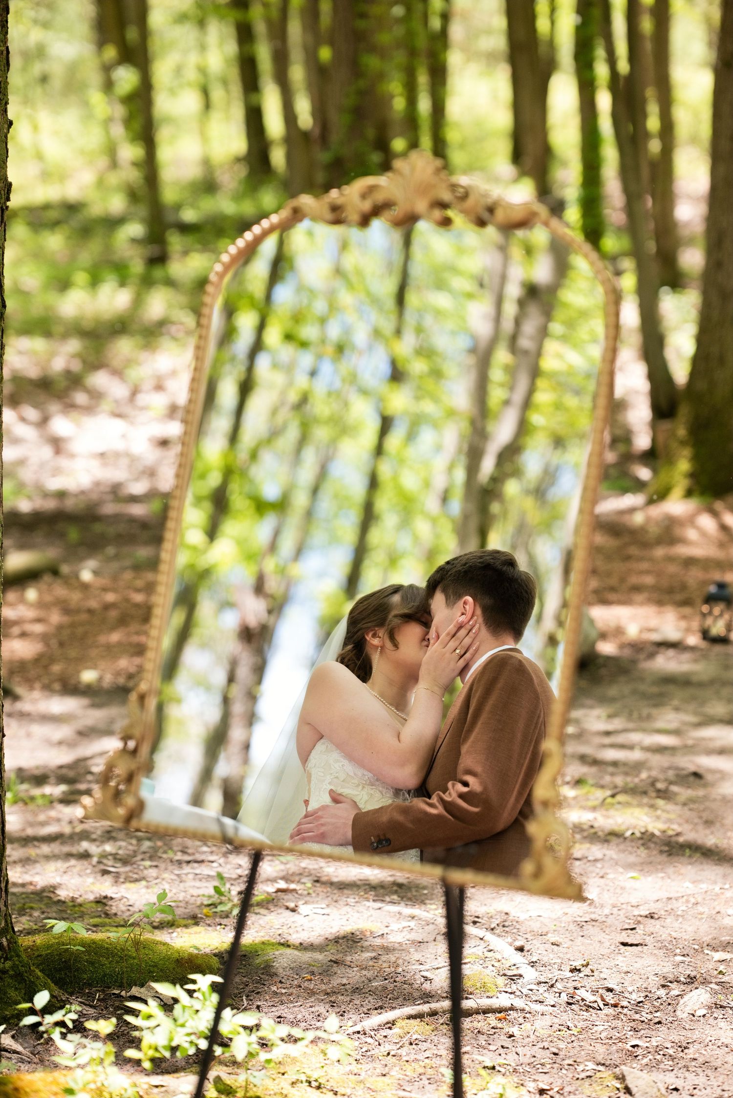 A couple shares a kiss reflected in an ornate gold mirror placed in a sunlit forest during their wedding.