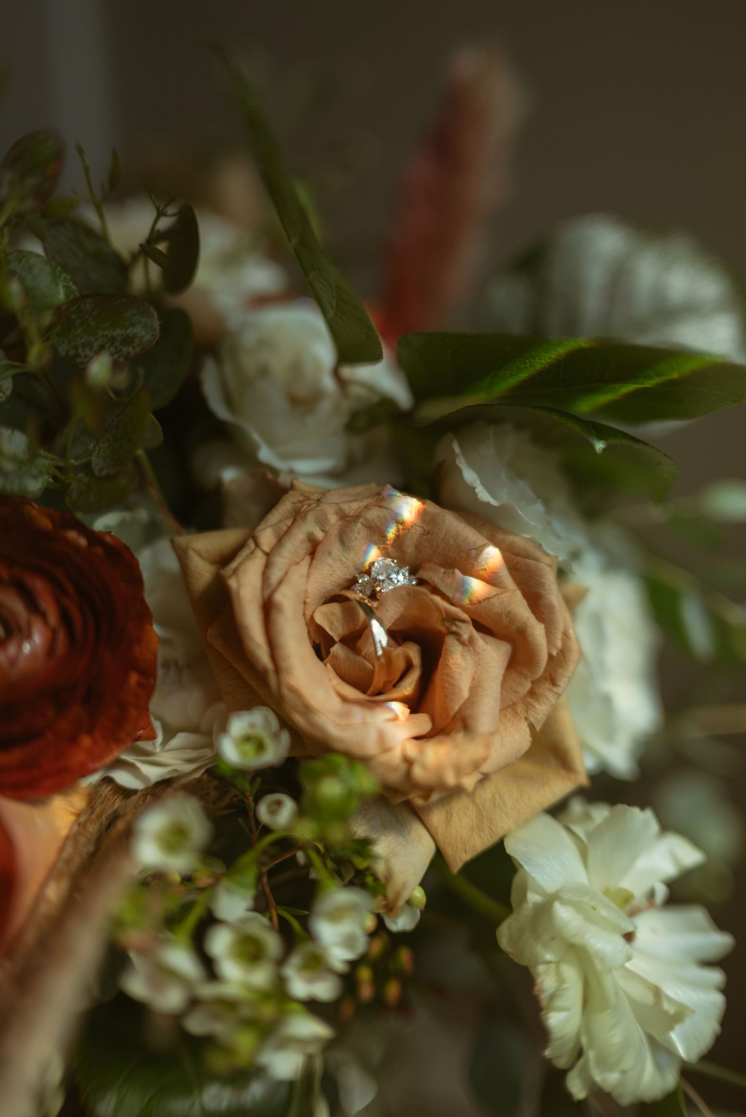 Diamond engagement ring nestled in a blooming tan rose surrounded by a lush bridal bouquet with red and white flowers.