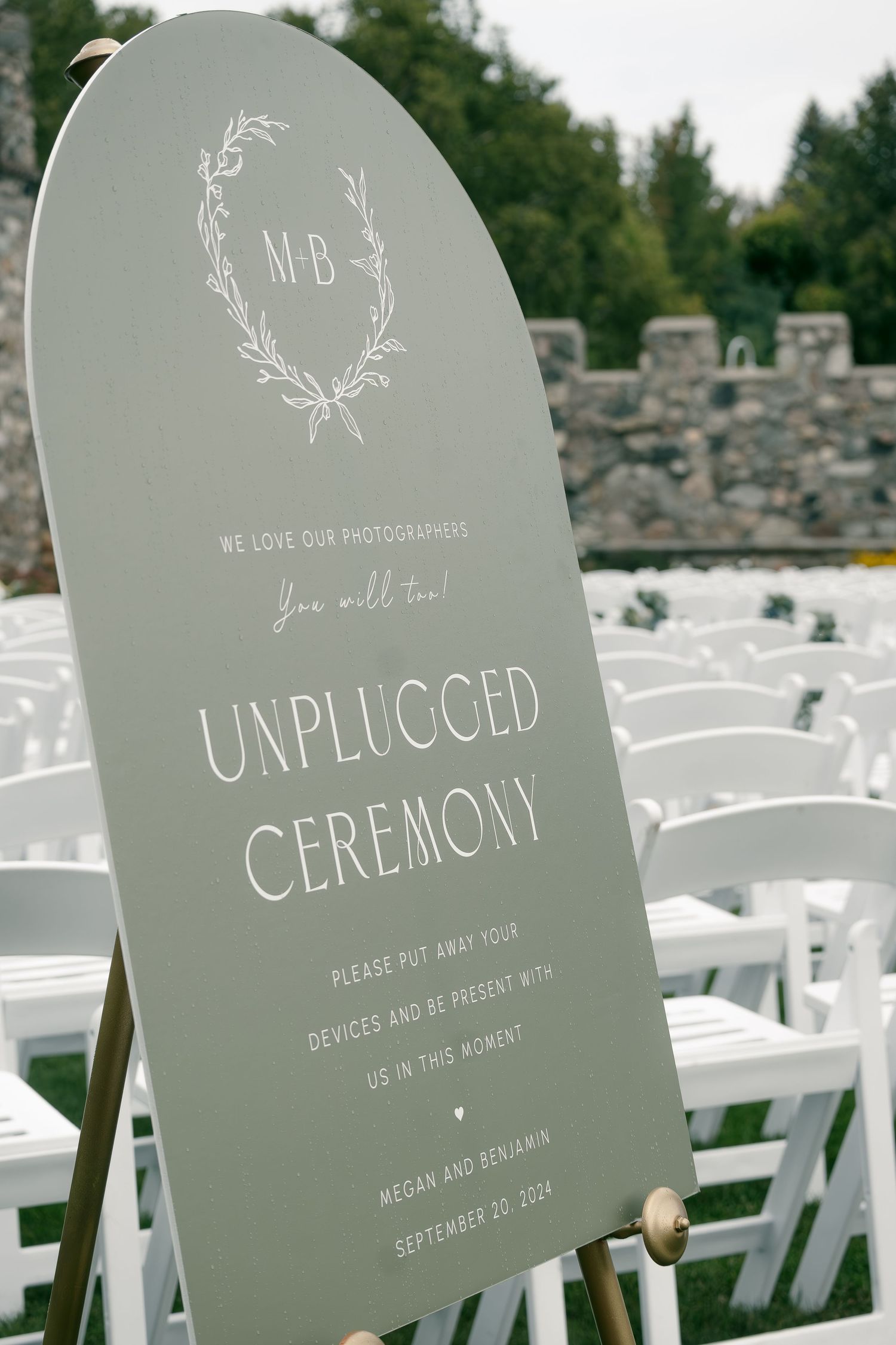 Gray wedding sign for unplugged ceremony with monogram displayed in front of white chairs.